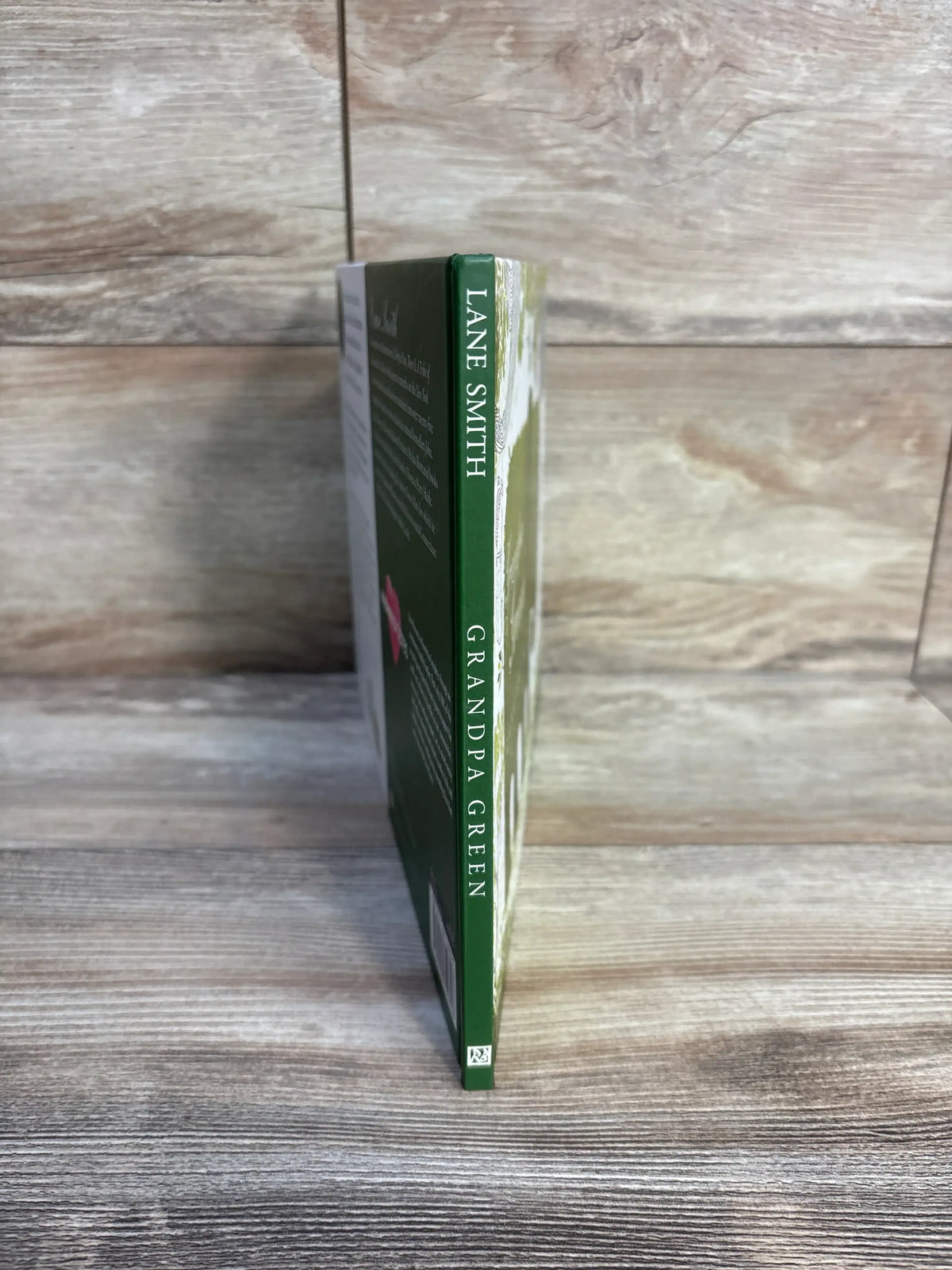 Side view of Grandpa Green hardcover book showcasing its green spine with white title and author text against a wood-grain background