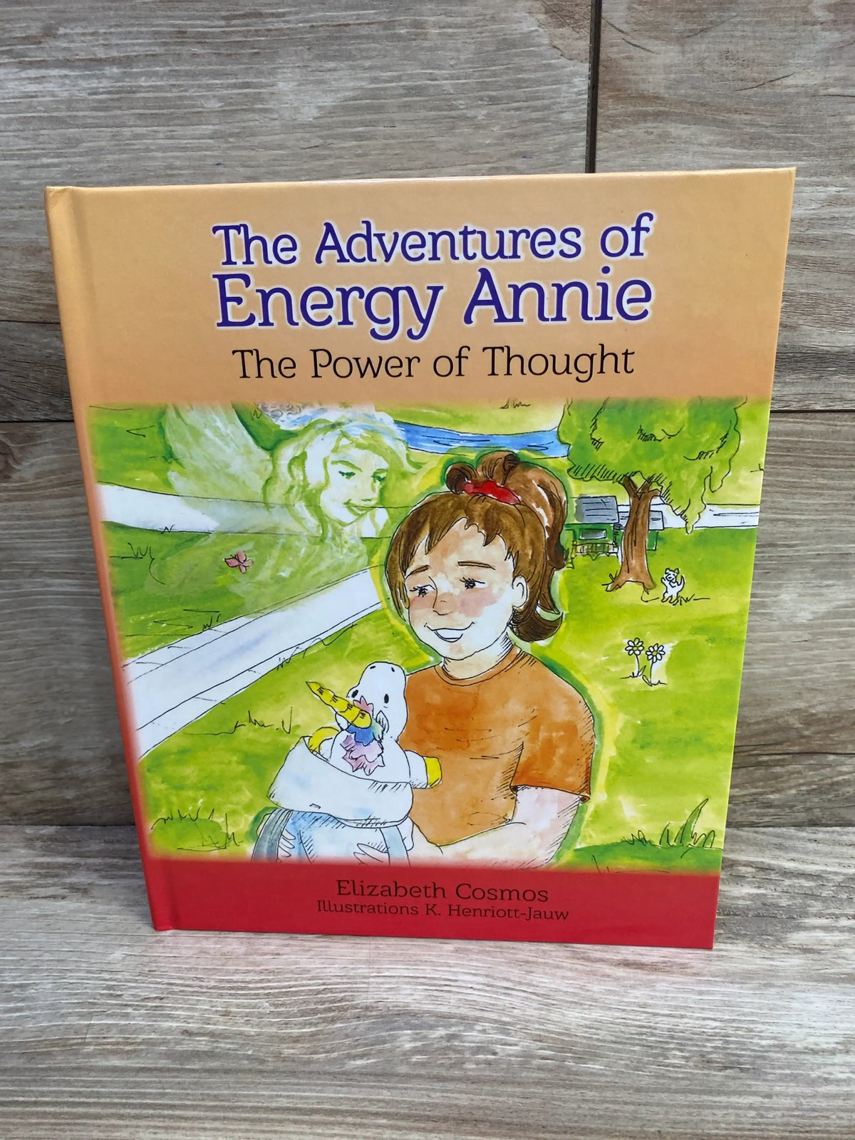 Front cover of hardcover book The Adventures of Energy Annie showing a girl holding a stuffed unicorn on a wood background