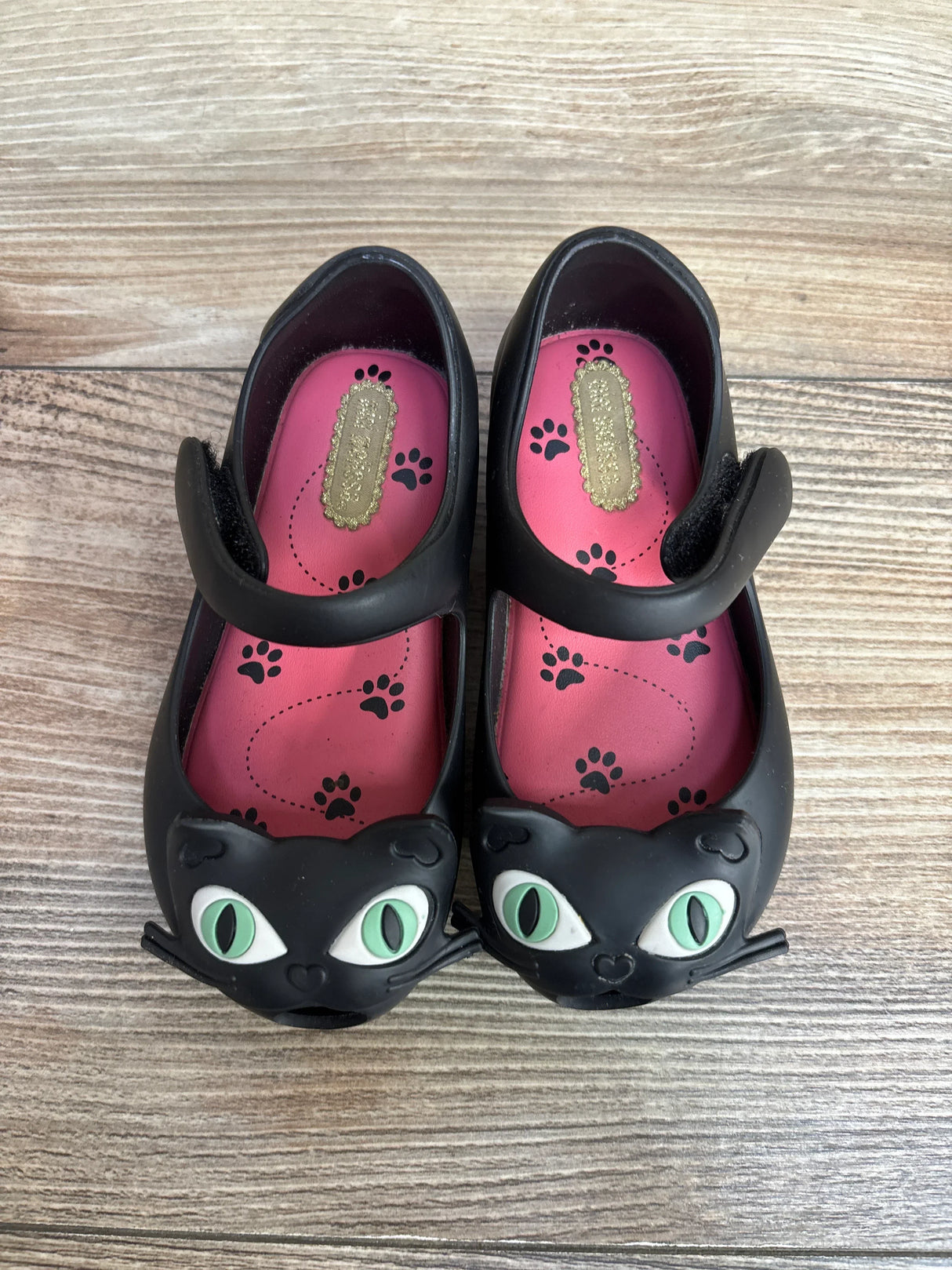 Top-down view of Mini Melissa Ultragirl II Cat Jelly flats in black showing cat-face toes, green eyes, and pink paw-print insoles on wood floor