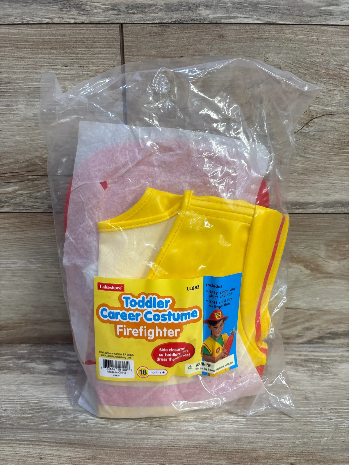 Front view of Lakeshore toddler firefighter costume in packaging with a yellow-and-red vinyl outfit, hat, and fire extinguisher