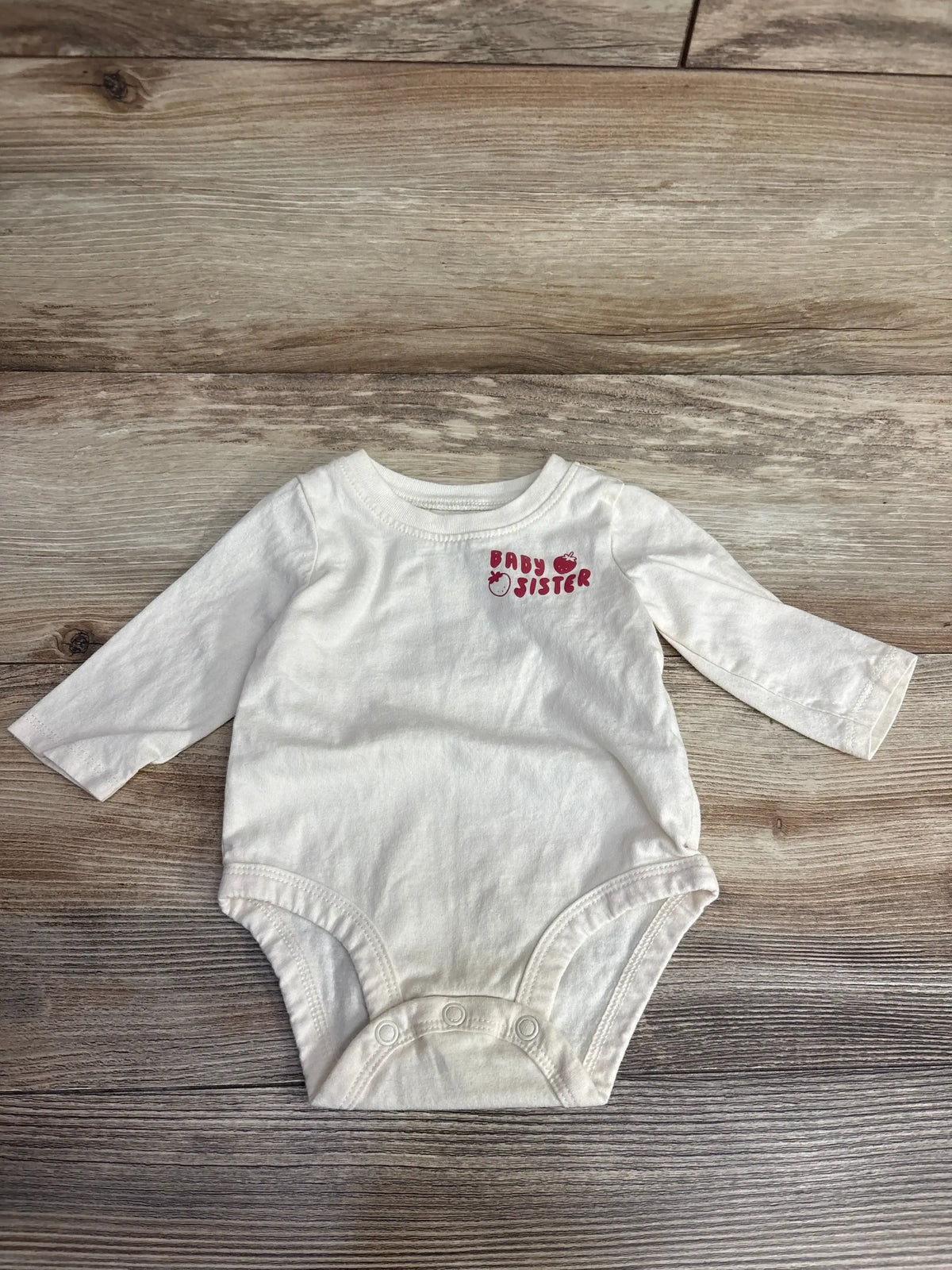 White long-sleeve baby onesie with red text and tomatoes reading 'Baby Sister' on wood background