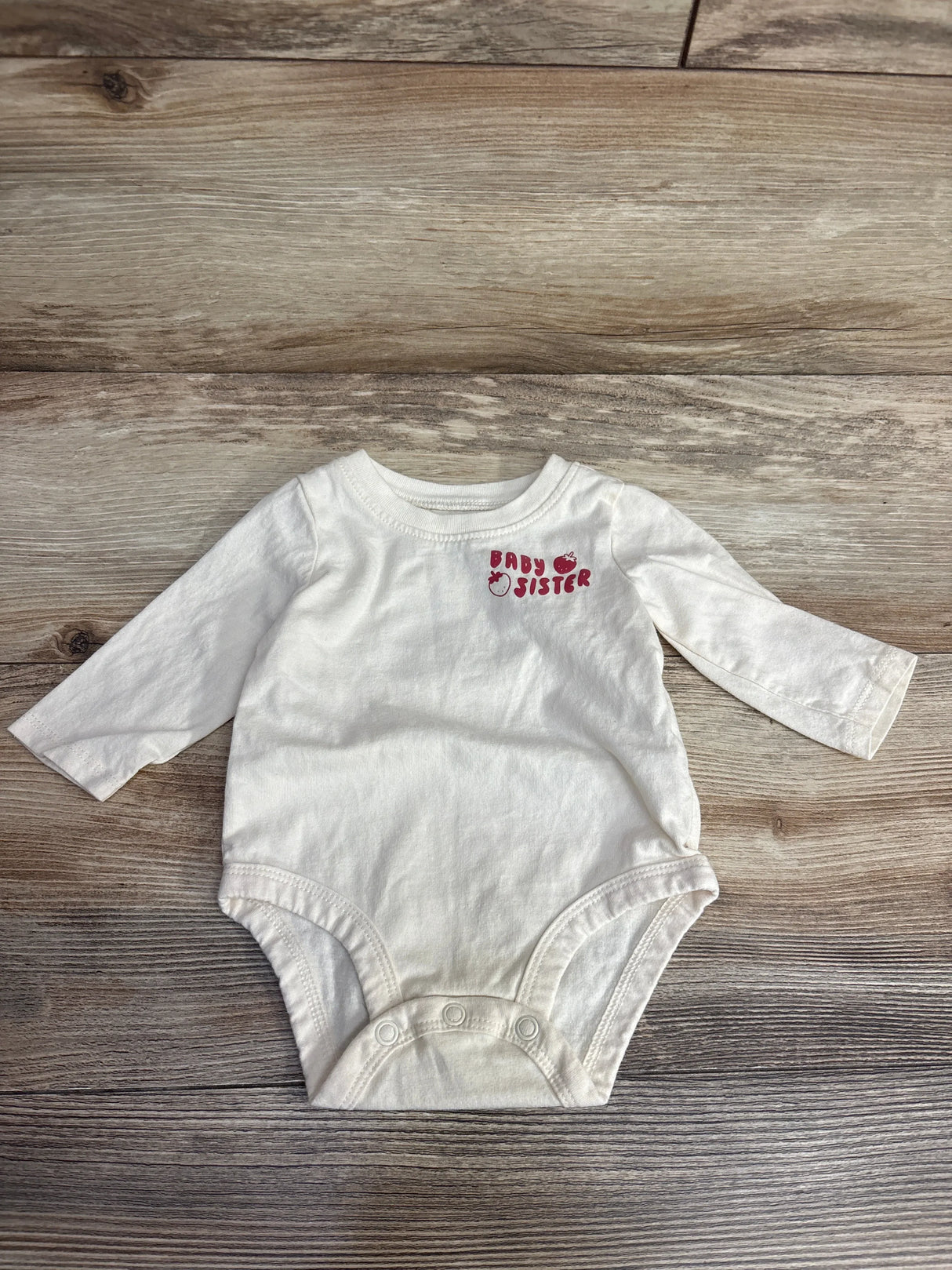 White long-sleeve baby onesie with red text and tomatoes reading 'Baby Sister' on wood background
