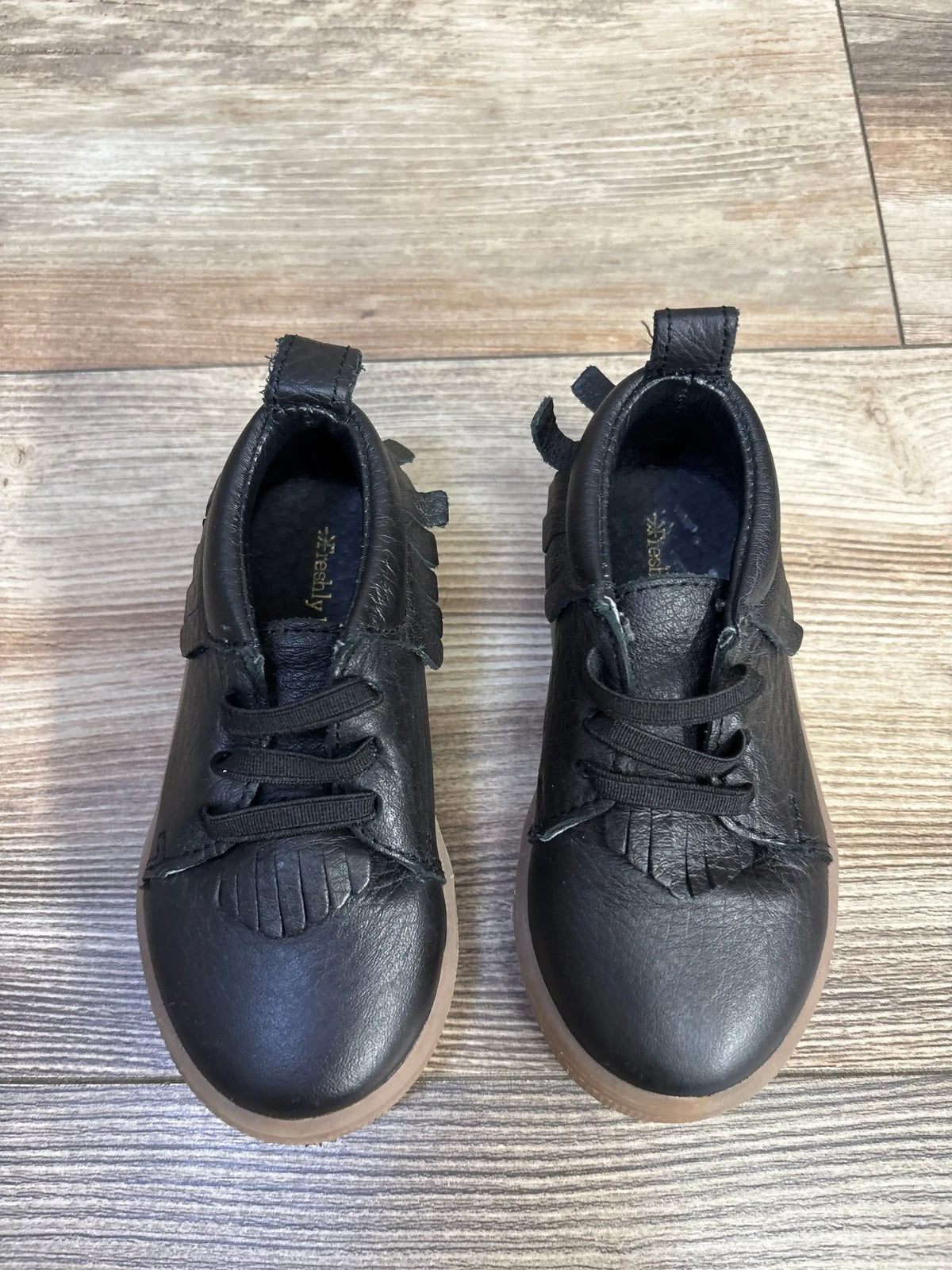 Black leather toddler shoes with fringed detail on wood floor