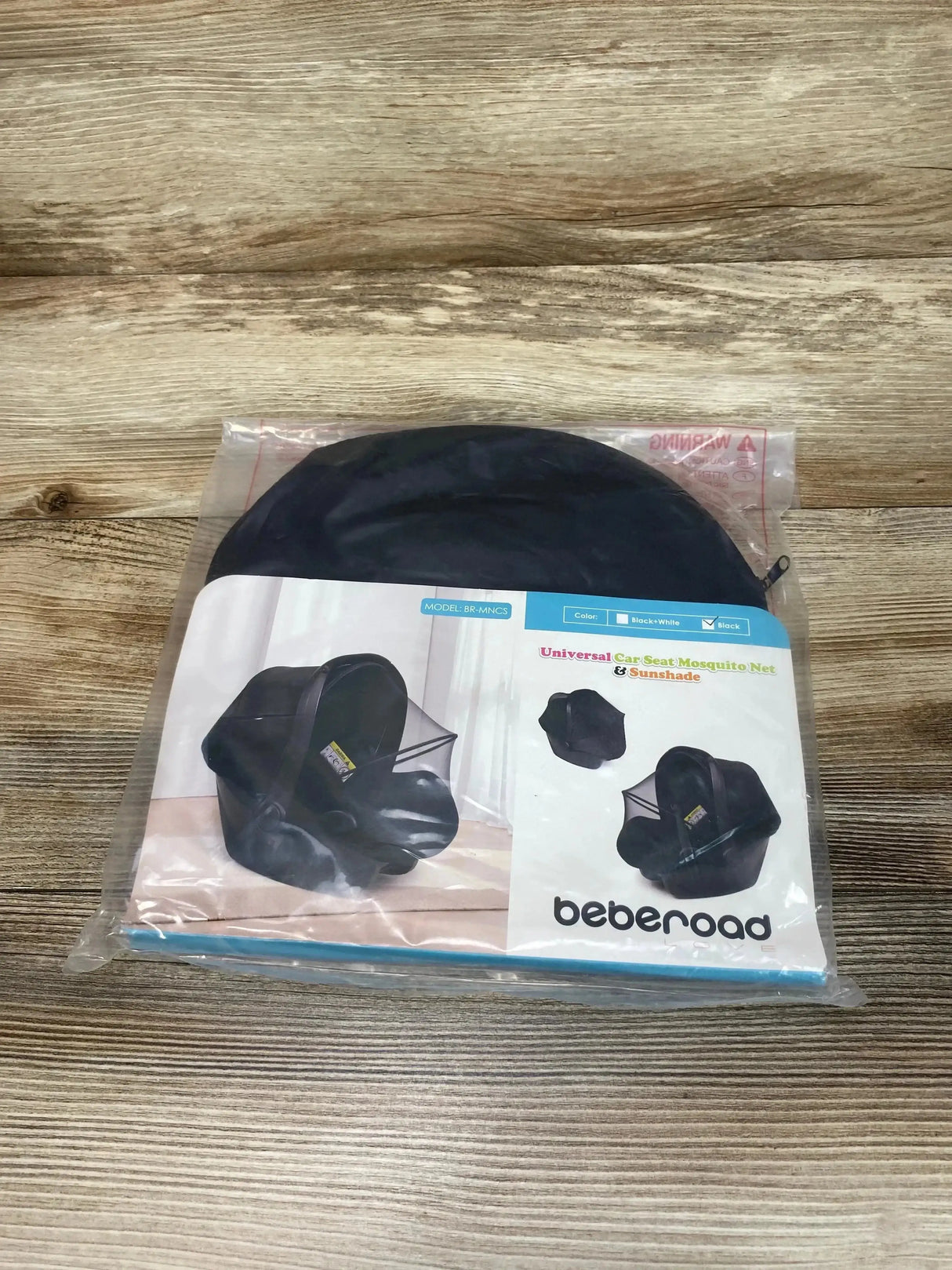 Top-down of packaged Beberoad universal car seat mosquito net and sunshade, folded black pop-up on wood background