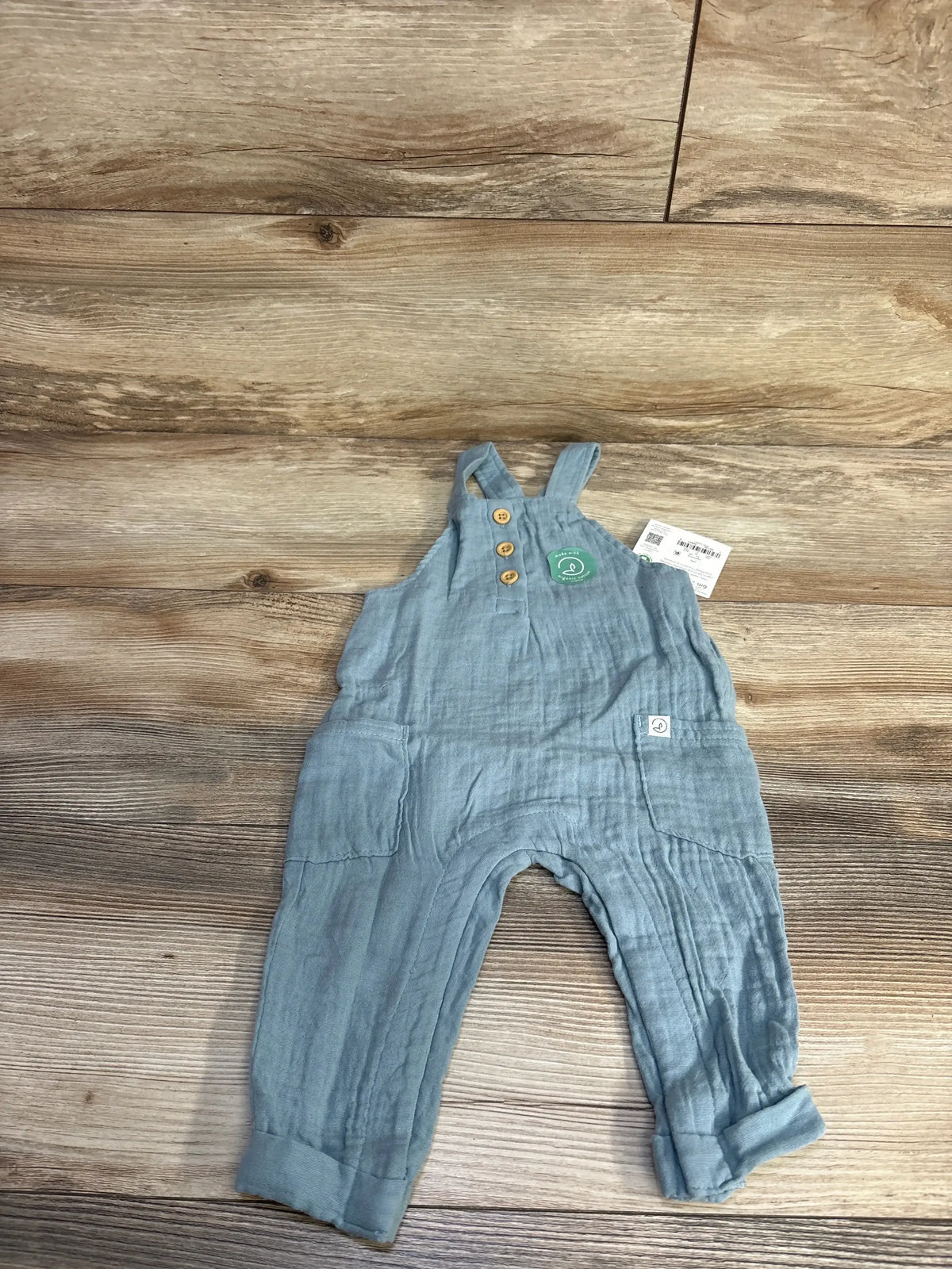 Flat lay of Little Planet blue gauze henley overalls 6m with wooden buttons, pockets and adjustable straps on wood floor