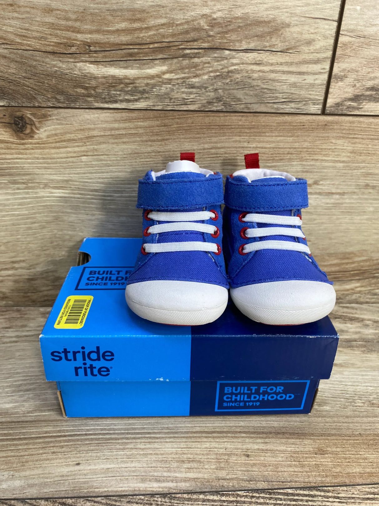 Front view of blue Stride Rite Soft Motion Danny toddler sneakers showcasing white rubber toe caps, elastic laces and Velcro ankle straps on a blue Stride Rite shoebox with wood backdrop