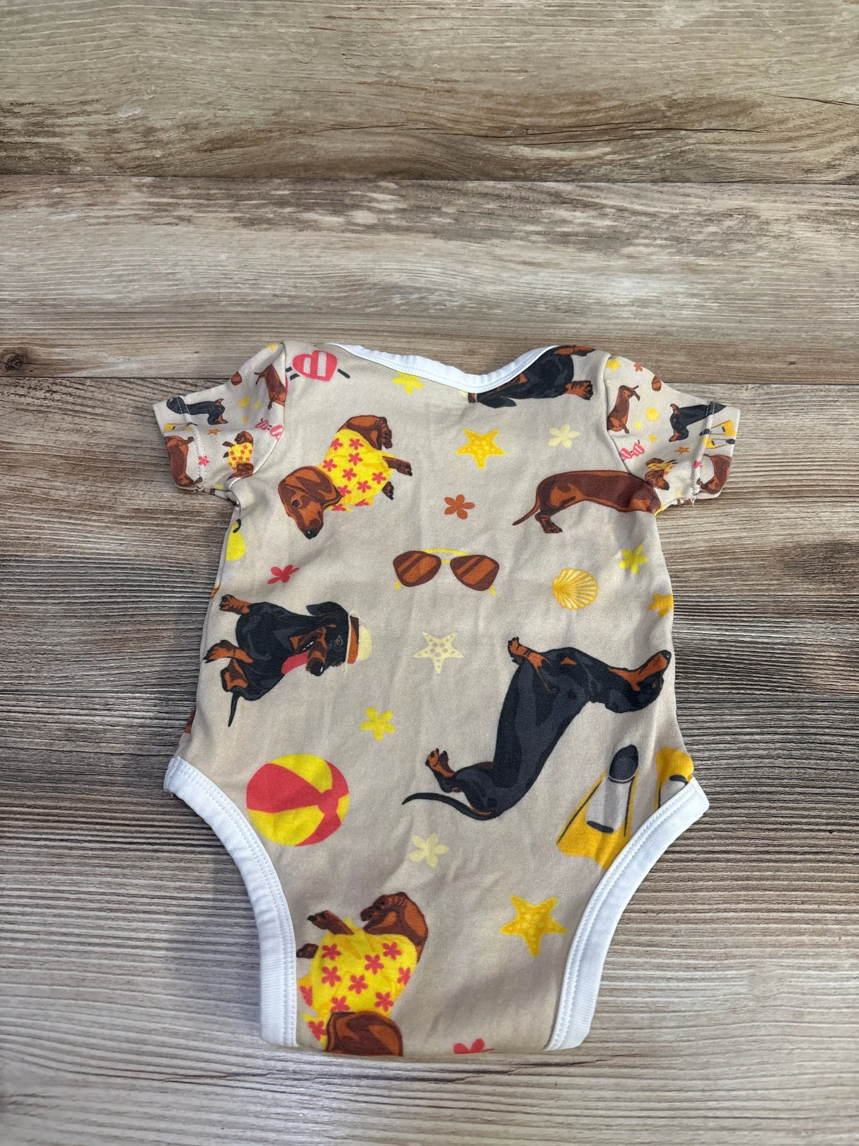 Top-down view of tan baby bodysuit showcasing dachshund dog print, short sleeves and white trim on wooden background