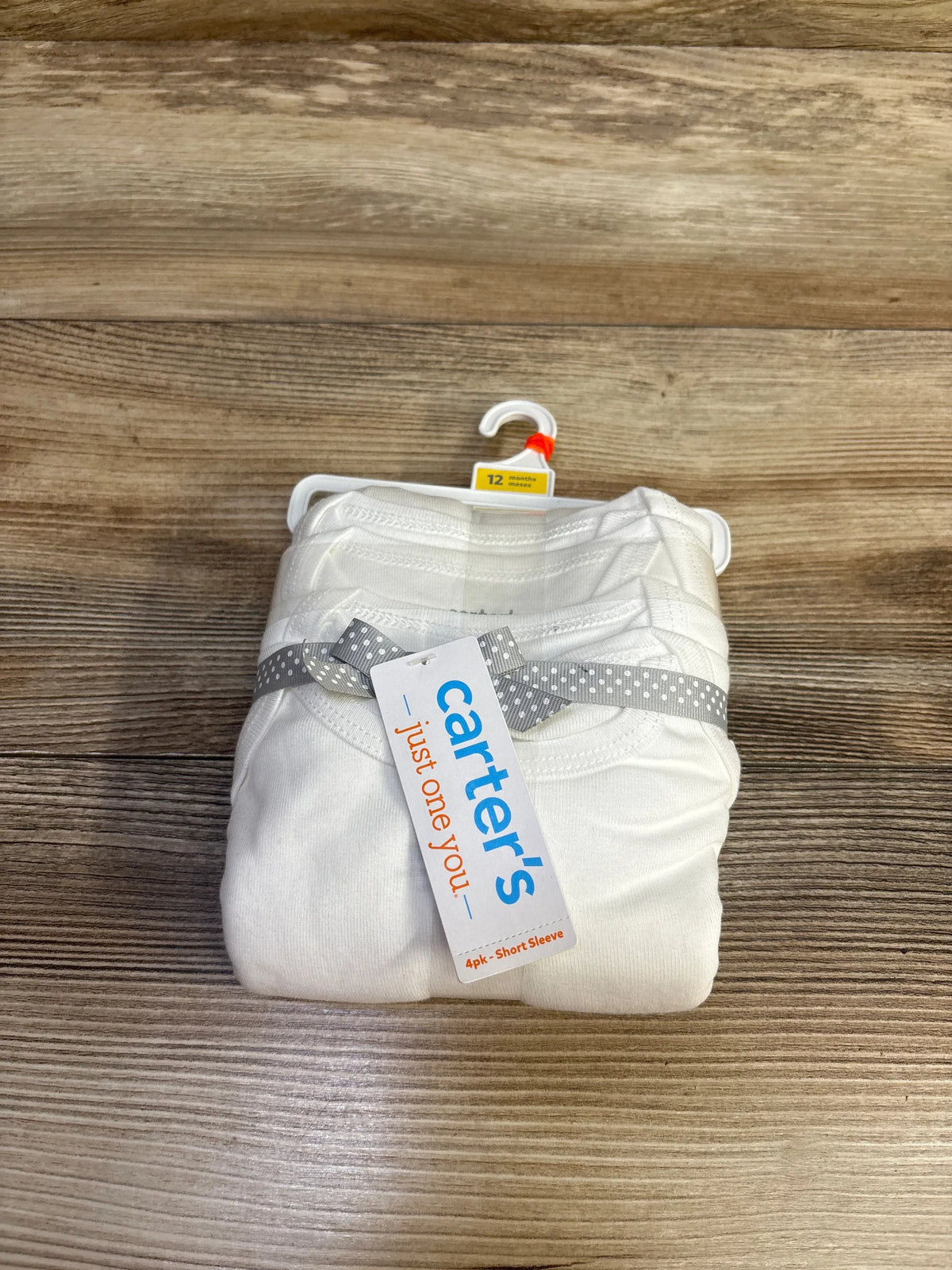 Top-down view of Carter’s 4-pack white short-sleeve baby bodysuits size 12 months showing brand tag on light wood background