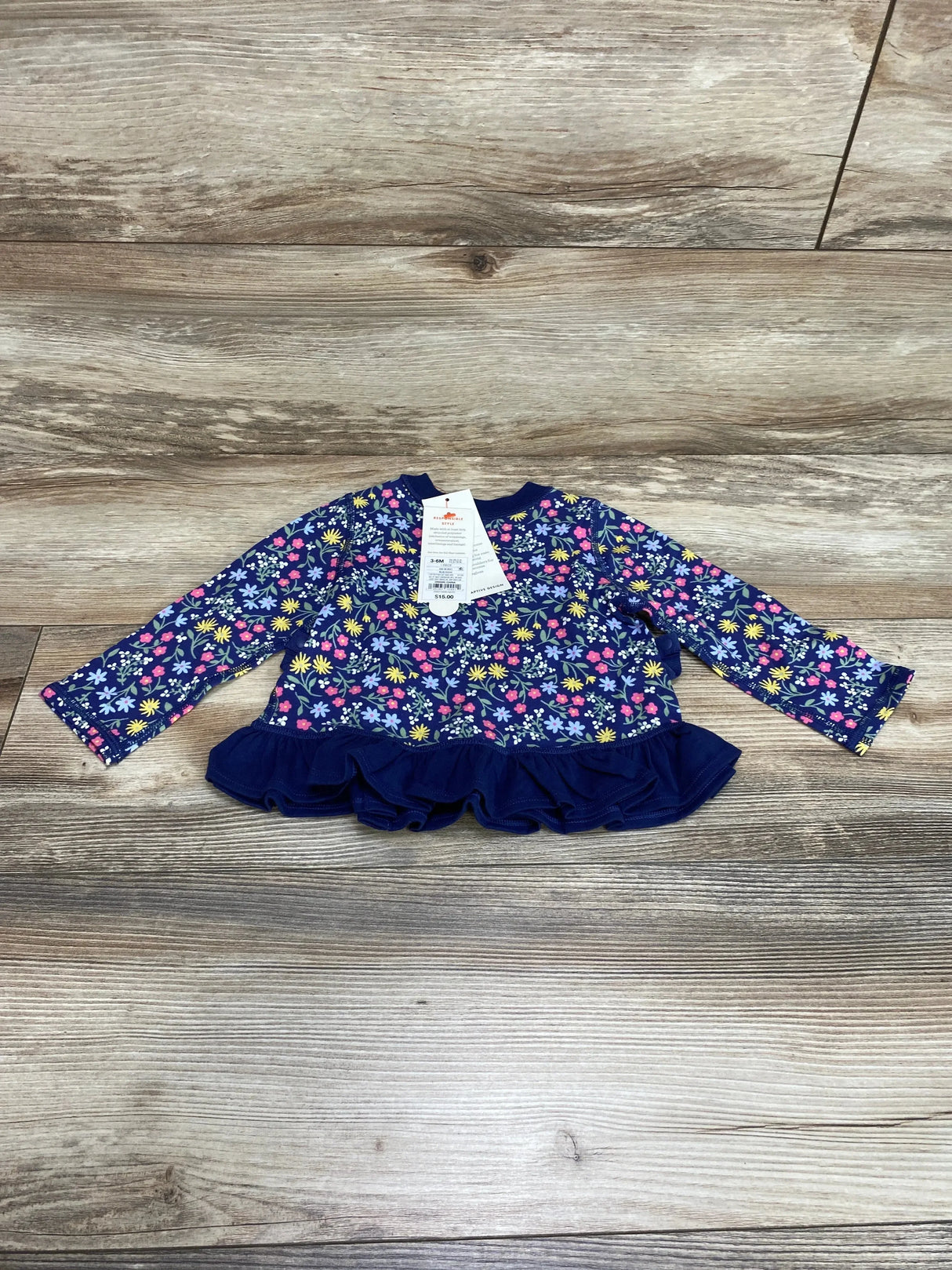 Front view of Cat & Jack navy floral long-sleeve peplum baby shirt showing ruffle hem on light wood floor background