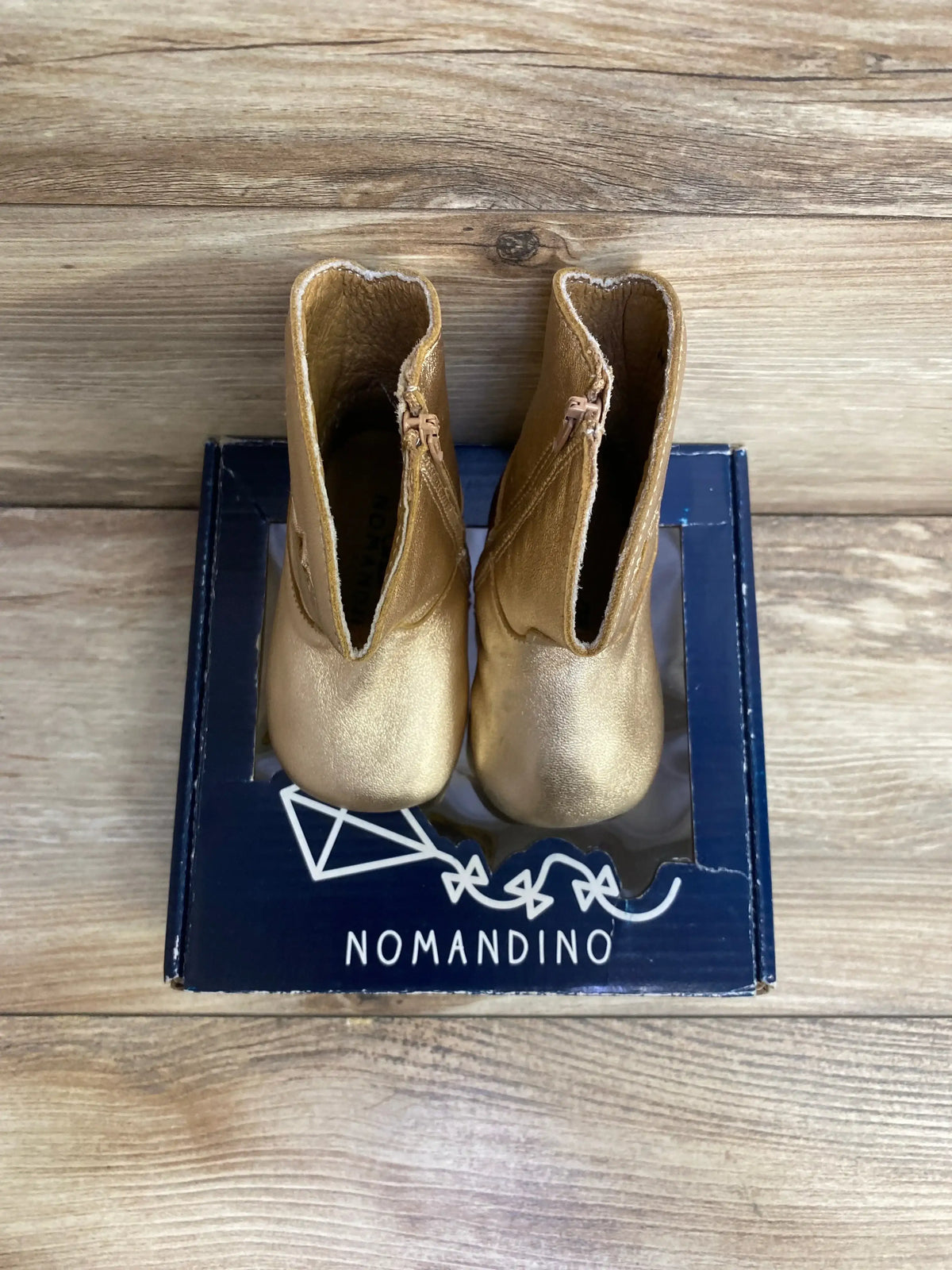Top view of Nomandino gold baby boots showing metallic finish and side zippers on navy Nomandino box with wood background