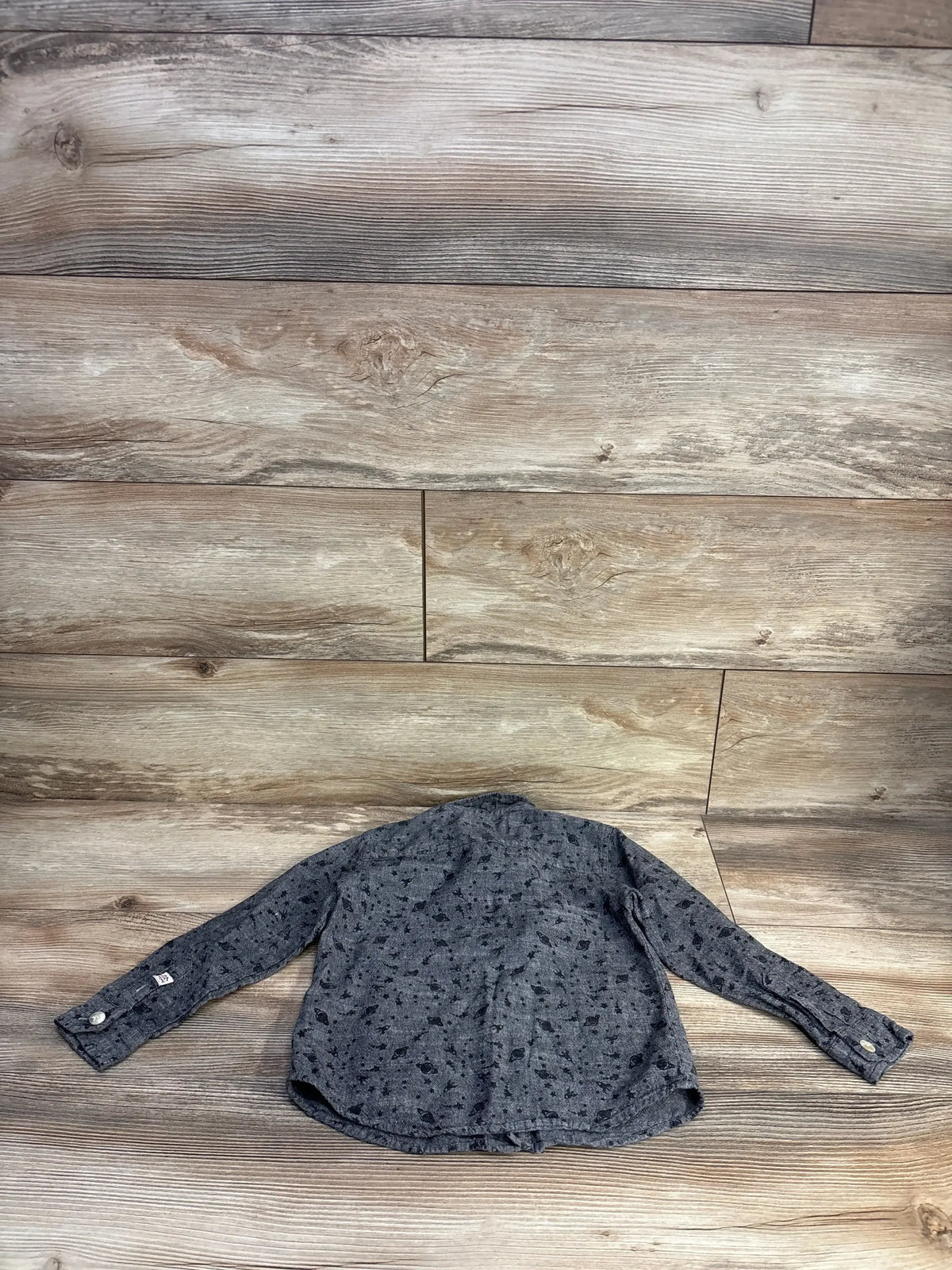 Back view of grey toddler button-up shirt with black space print, long sleeves and white buttons on wood plank background