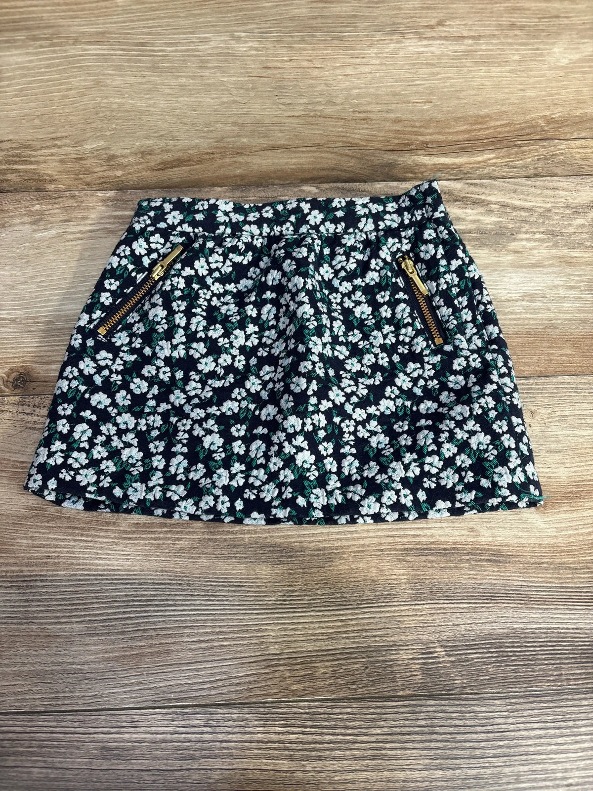 Flat lay of toddler black floral skirt with white and green flowers, gold zip pockets and elastic waist on wood background