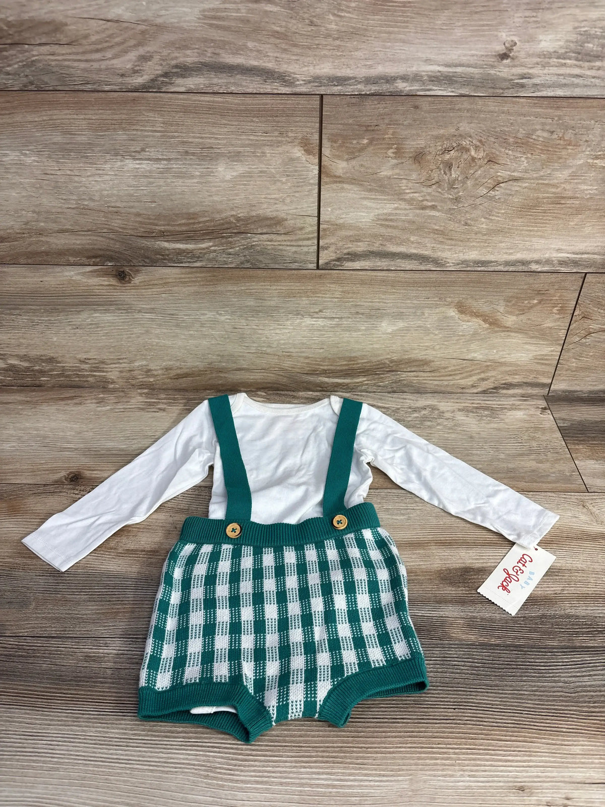 Baby outfit with white long sleeve top and green checkered knit suspender shorts on wooden floor