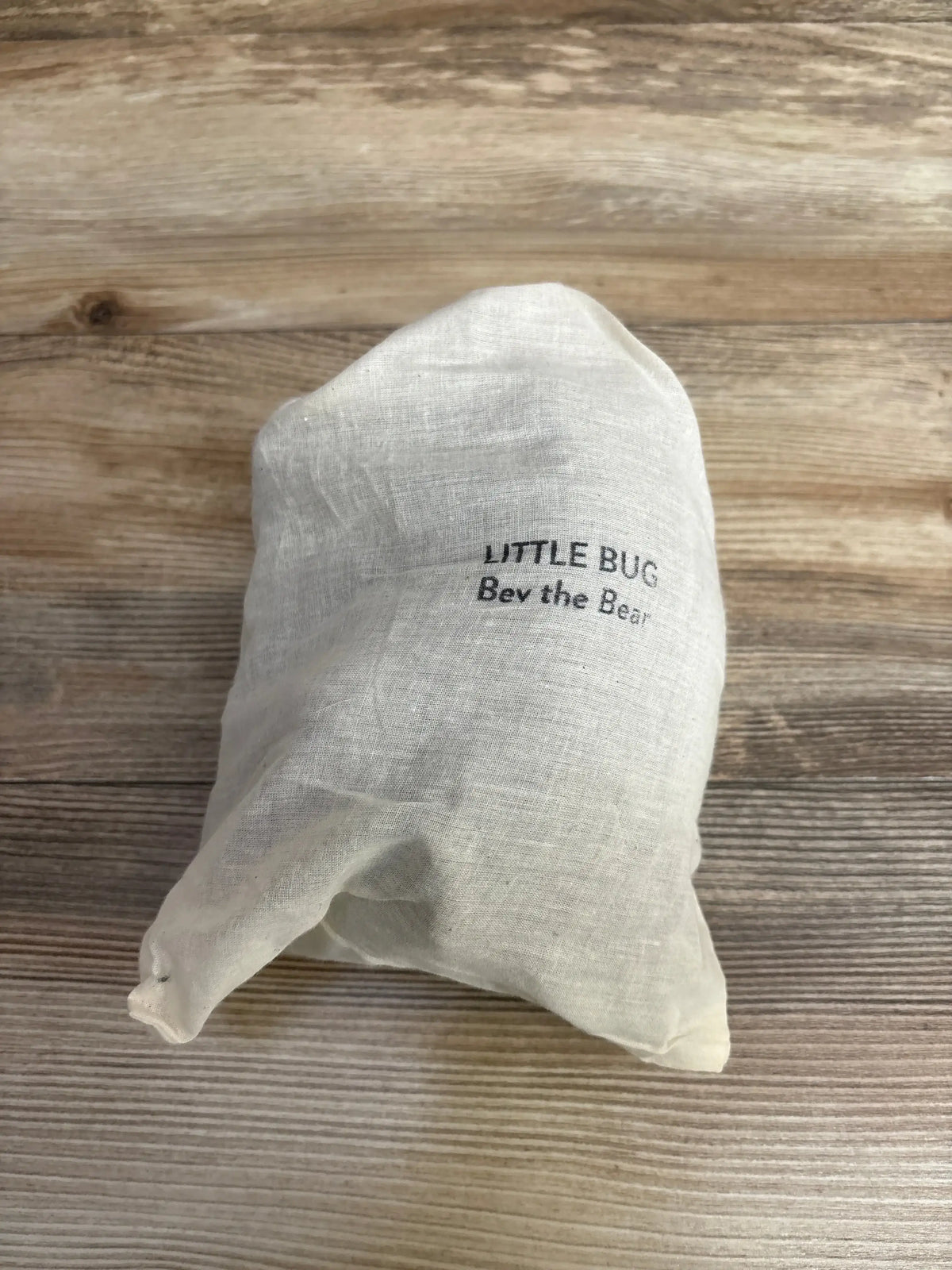 Packaged view of Little Bug Bev the Bear lovie inside white linen drawstring bag labeled LITTLE BUG Bev the Bear on wooden surface
