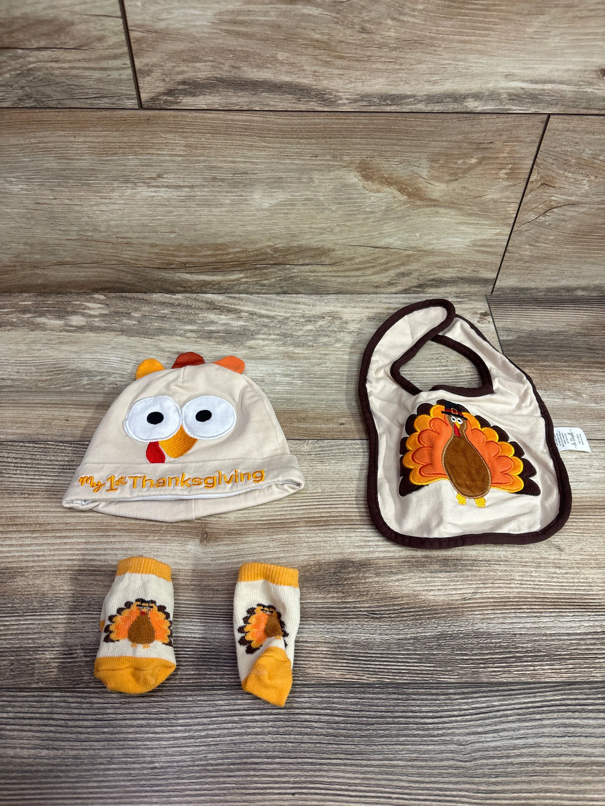 Top view of beige baby Thanksgiving hat bib and socks set with turkey designs and My 1st Thanksgiving text on wood floor