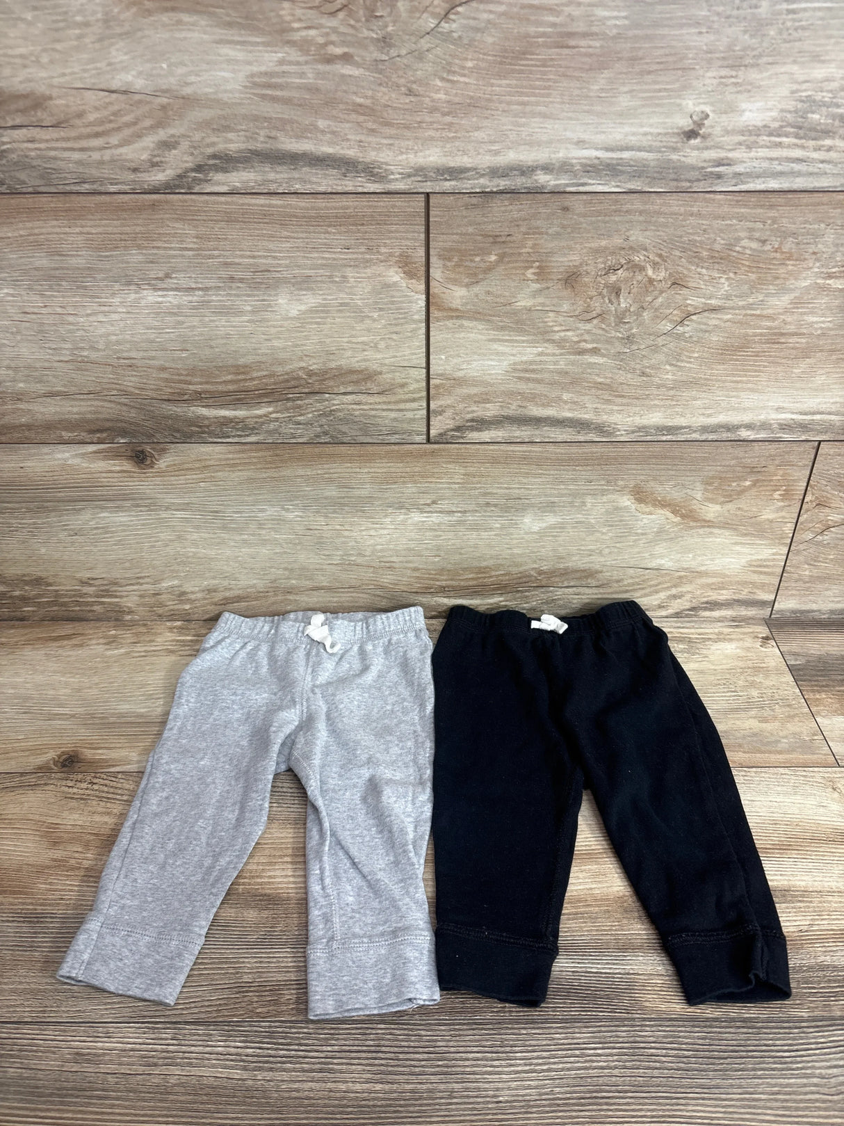 Flat lay of two Carter's baby pull-on pants, black and gray, showing elastic waist with white ties and cuffed legs on wood floor