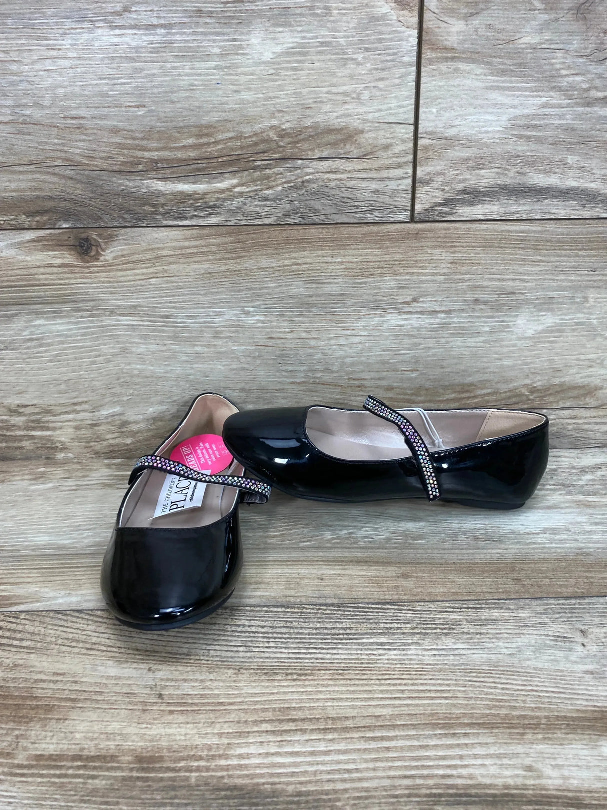 Three quarter view of girls' black faux patent leather ballet flats showing jeweled Mary Jane strap on a wood floor background