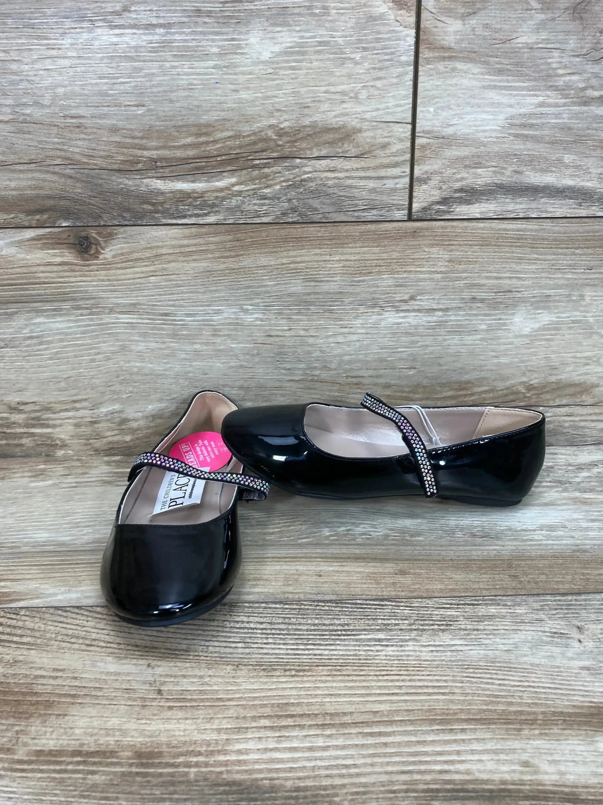 Three-quarter view of Children’s Place faux patent ballet flats in black showcasing rhinestone strap and pink hangtag on wood plank floor