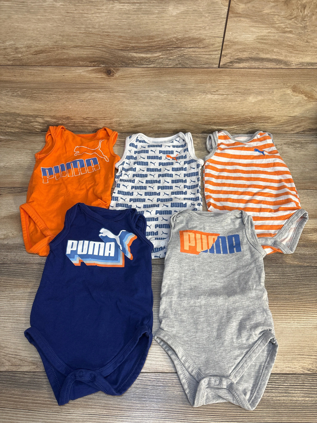 Assorted Puma baby bodysuits in orange, blue, and gray on wooden floor
