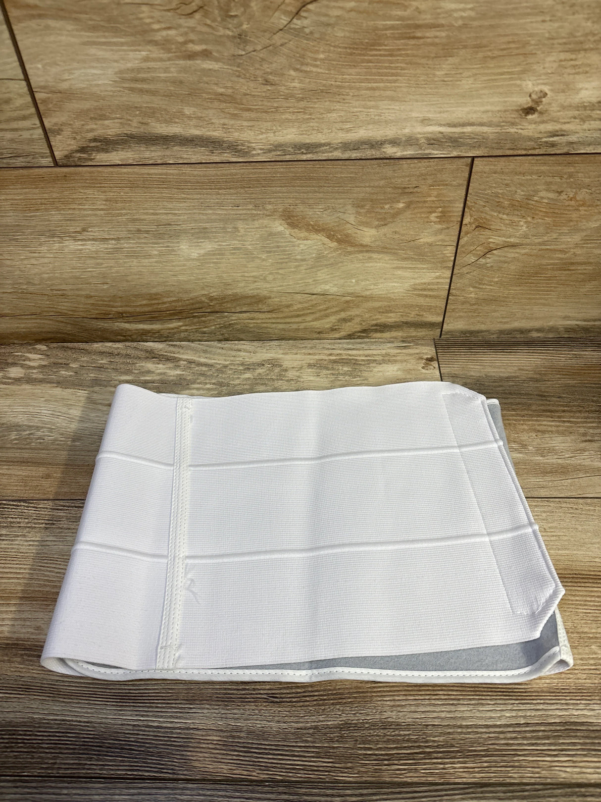 Top-down view of 9-inch BREG abdominal binder in white showcasing horizontal support bands and hook-and-loop closure with wood plank background