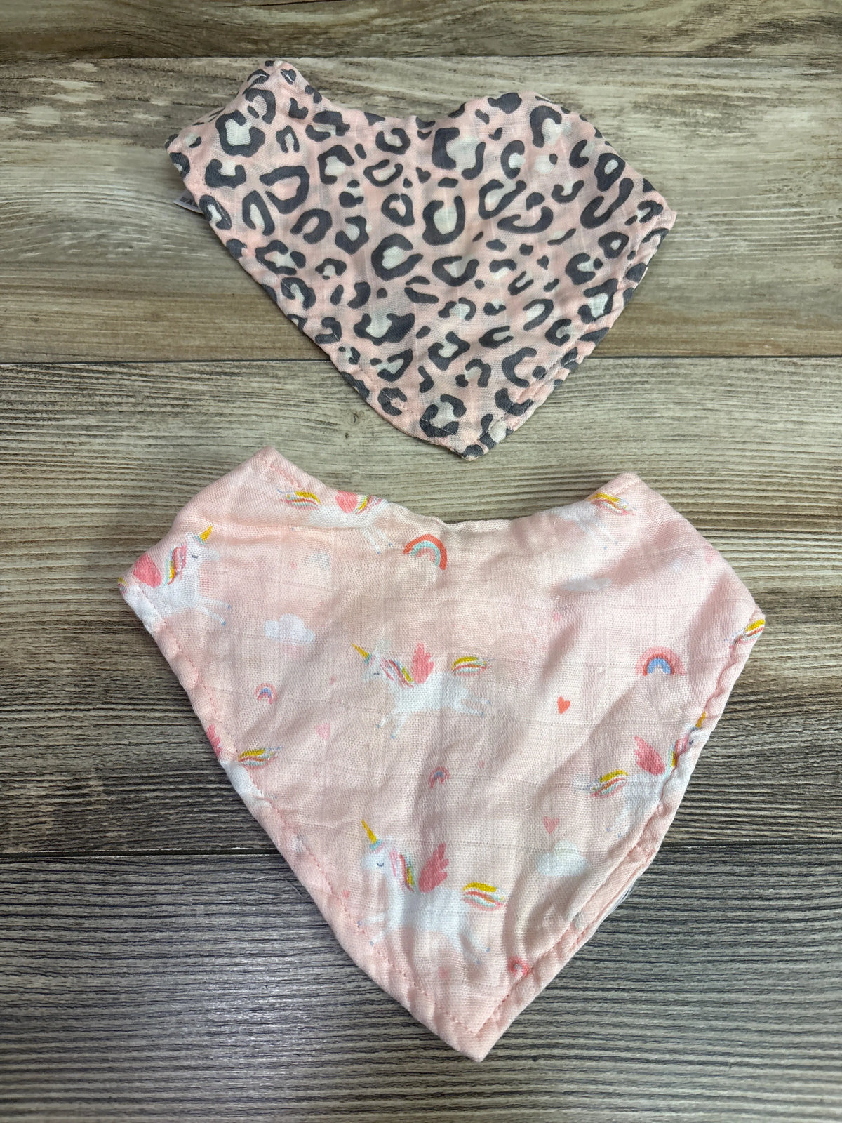 Top-down view of Angel Dear 2pk bandana bibs showcasing pink leopard and rainbow prints on a wood-grain background