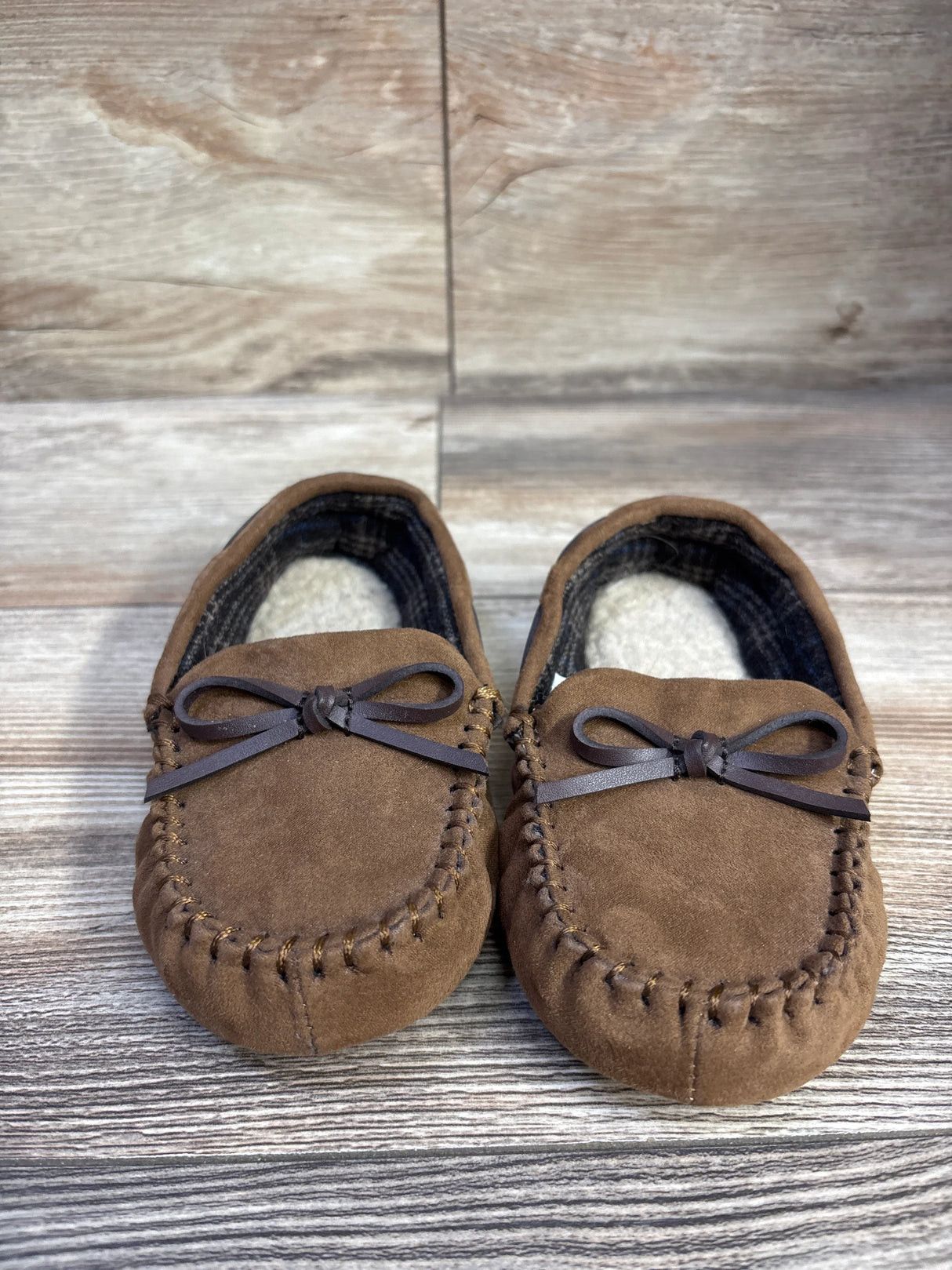 Front view of Cat and Jack Lionel brown moccasin slippers with stitched toe, bow detail and soft faux fur lining on wood floor
