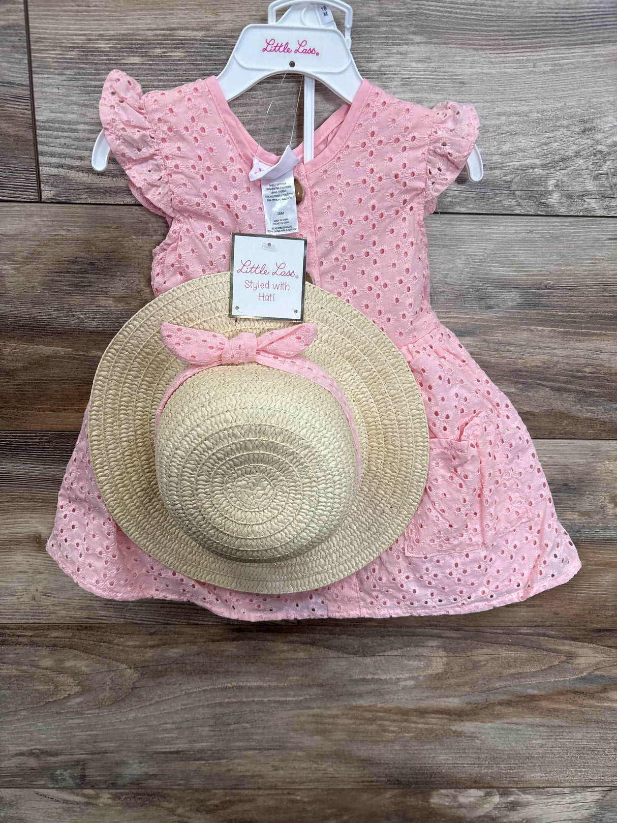 Pink eyelet baby dress with matching straw sun hat, displayed on wooden background.