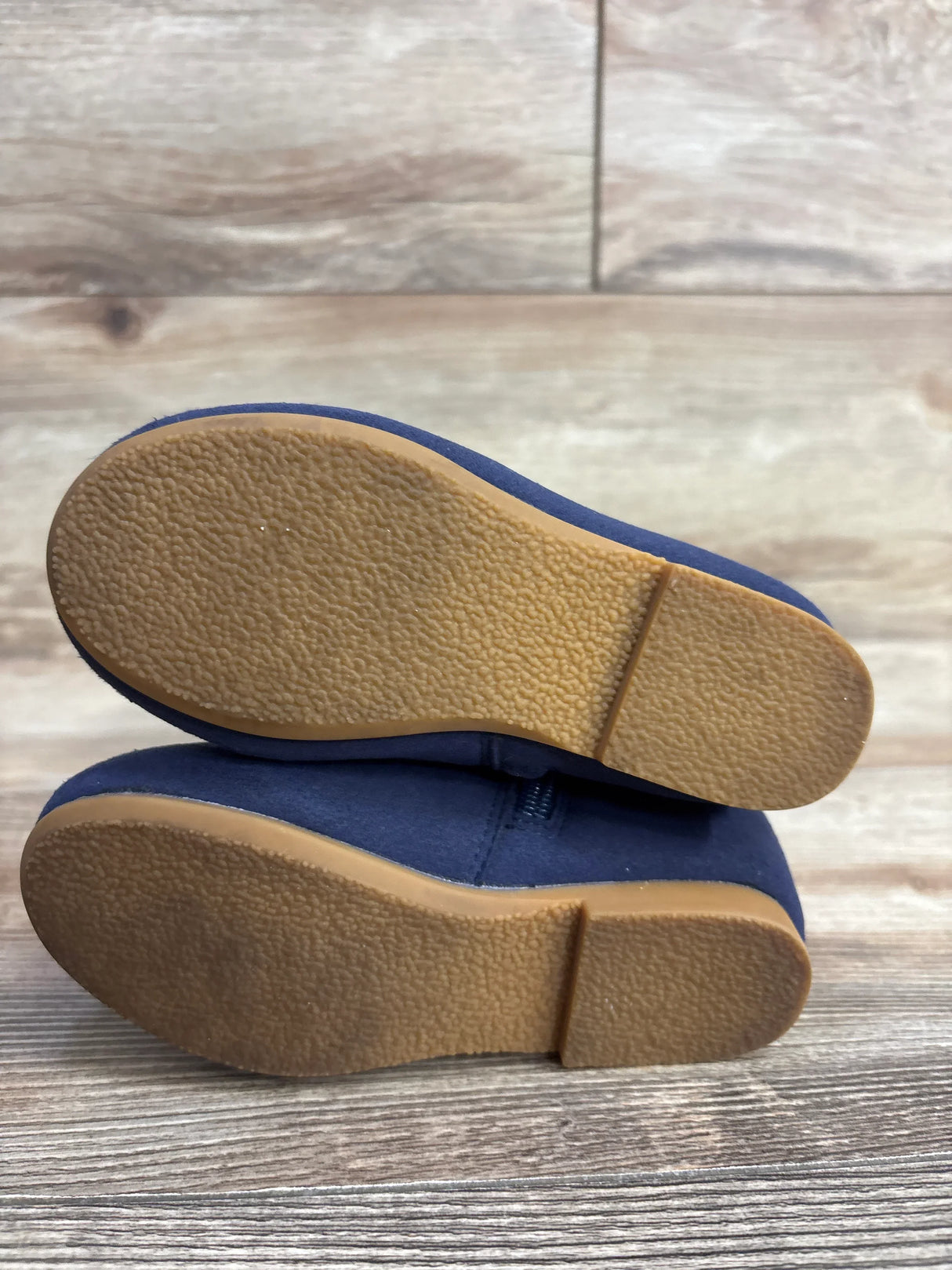 Bottom view of pair of navy blue suede toddler shoes showing tan rubber outsoles with textured tread on wood plank floor