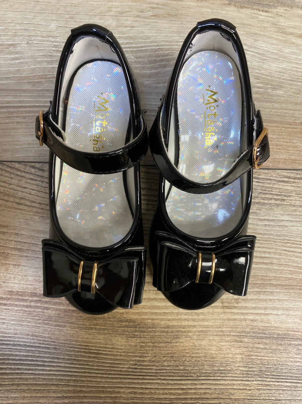 Girls' black patent leather dress shoes with bows and gold buckles on wood floor