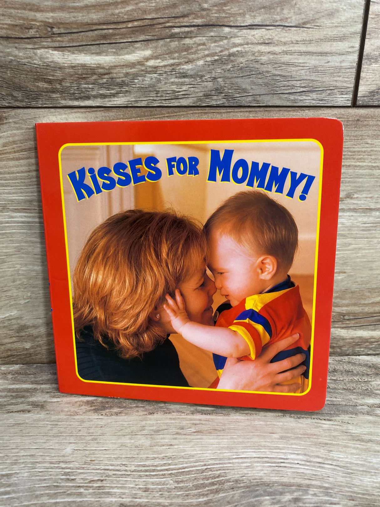 Front cover of board book Kisses for Mommy! showing mother and toddler touching noses, red border, wood background