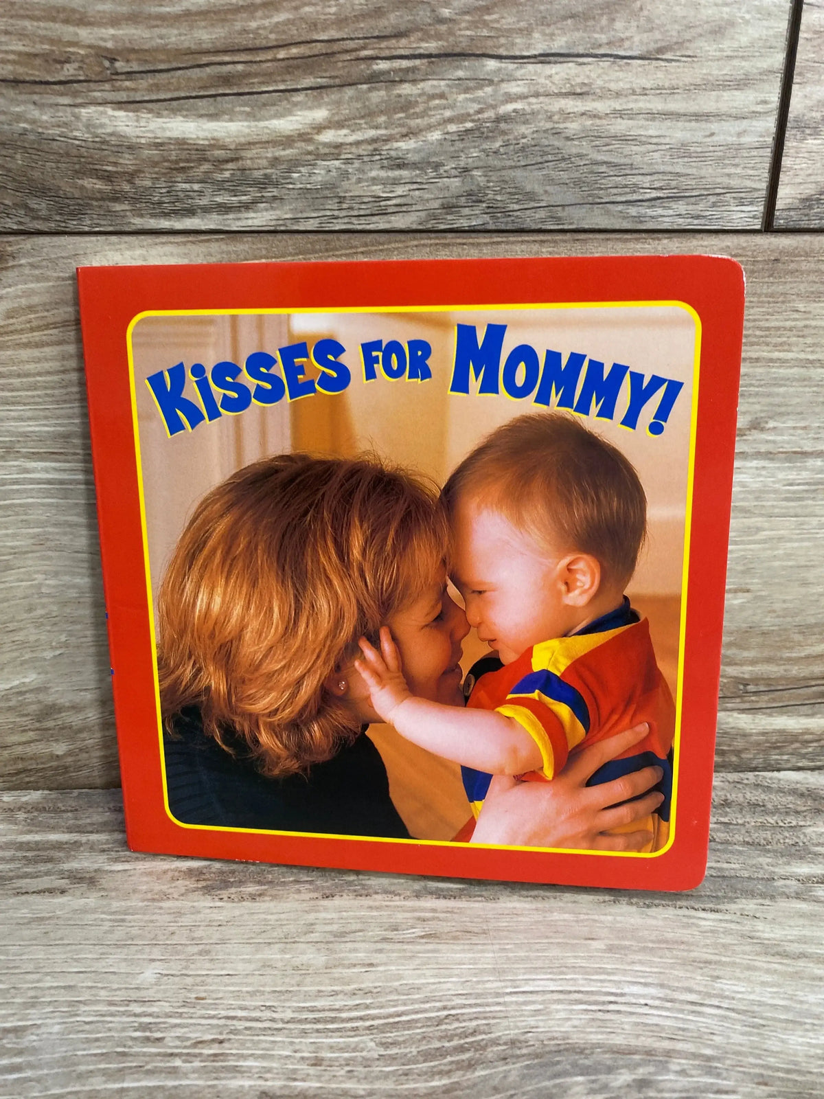 Front cover of board book Kisses for Mommy! showing mother and toddler touching noses, red border, wood background