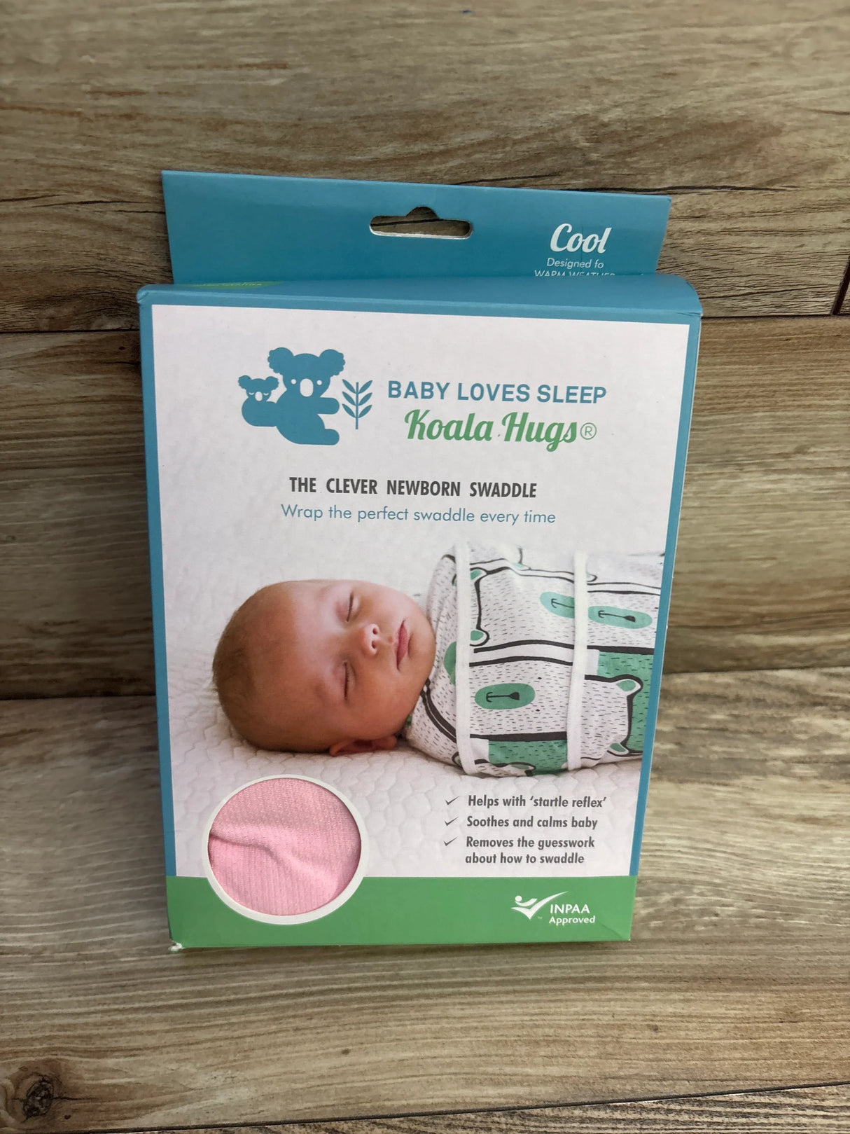 Front view of NEW Baby Loves Sleep Koala Hugs Newborn Swaddle in original retail box showing pink fabric swatch and sleeping baby graphic on wood plank floor