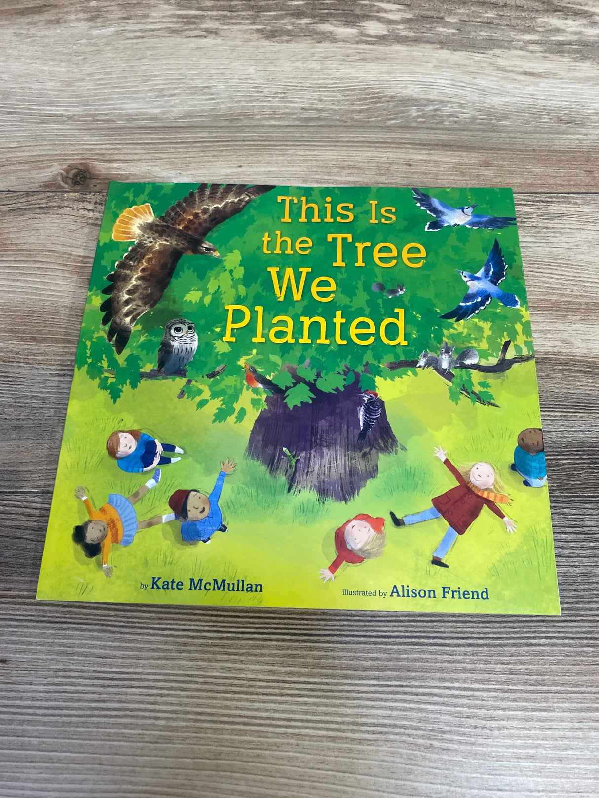 Front view of paperback This Is the Tree We Planted showing illustrated cover with tree, children, and birds on wood tabletop