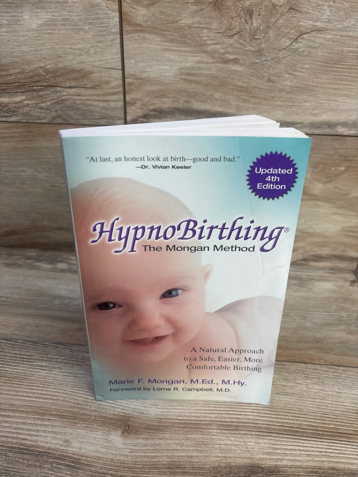 Front cover of HypnoBirthing paperback book showcasing a smiling baby and updated fourth edition badge on a wood surface