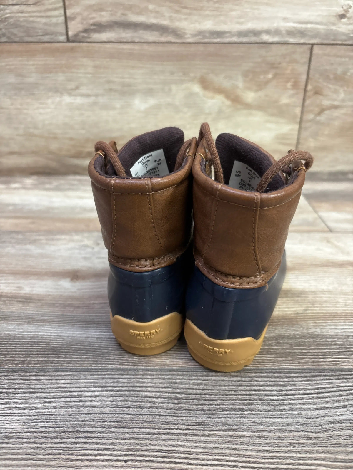 Rear view of Sperry Port Alpine waterproof duck boots in brown and navy with tan soles on a wood floor