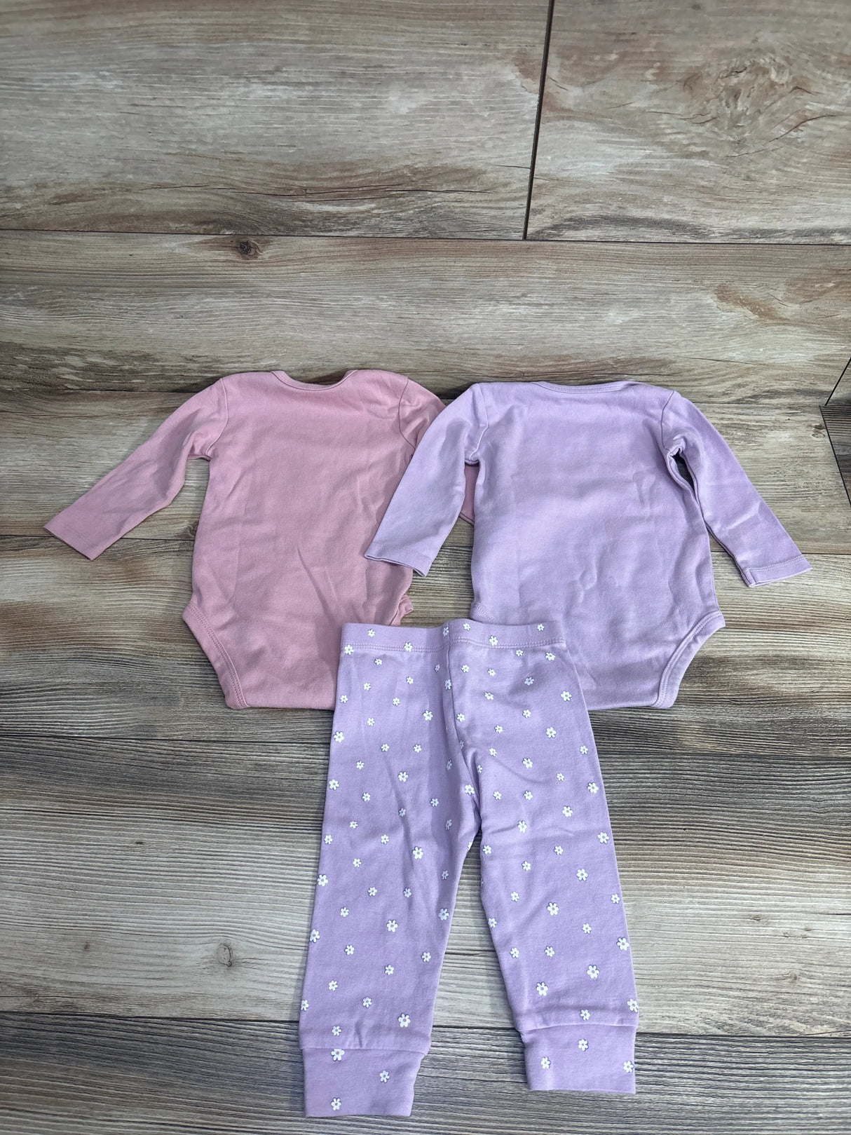 Flat lay of baby girl 3pc set with pink and lavender long-sleeve bodysuits and matching lavender floral print pants on wood plank floor