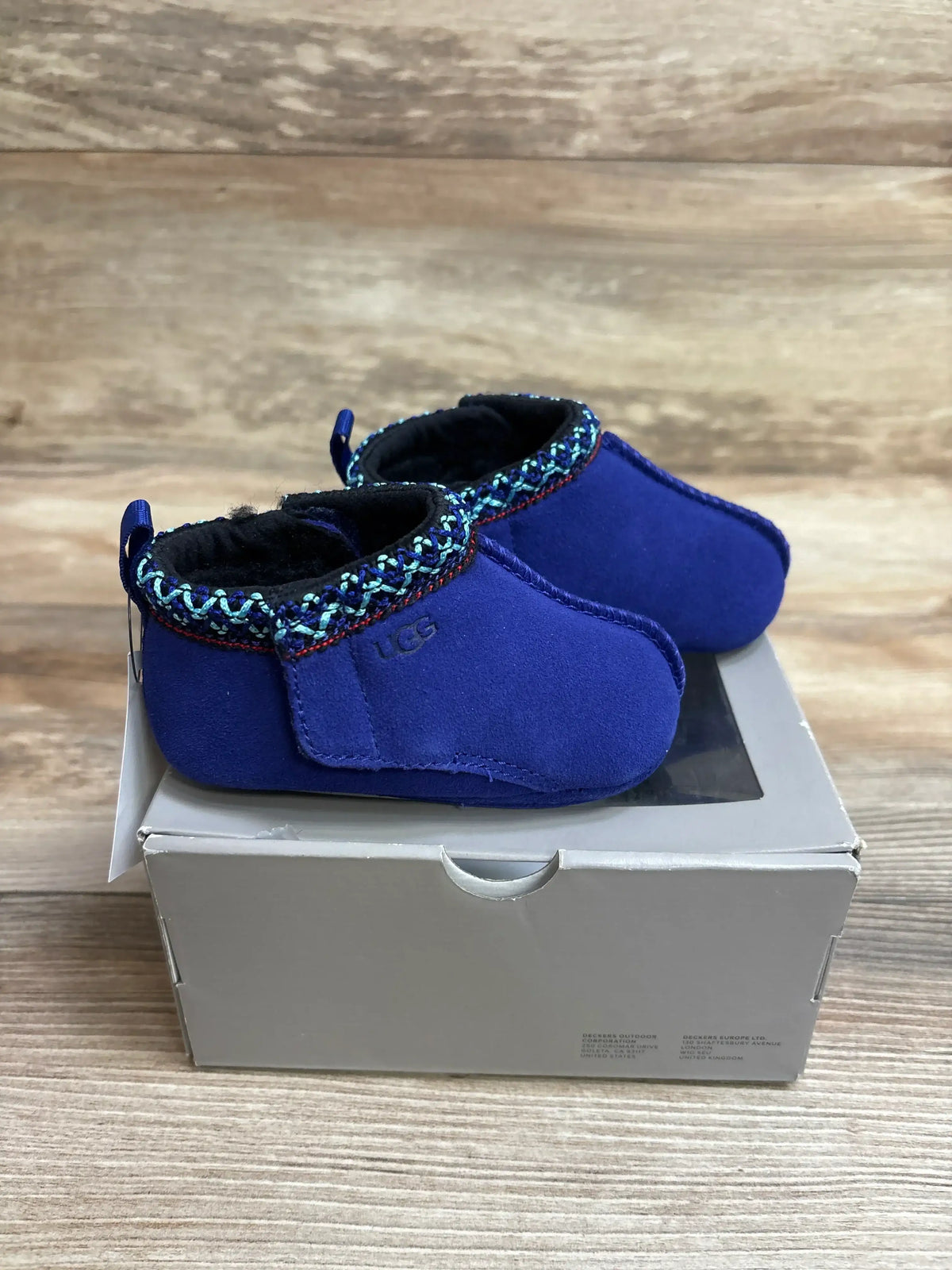 Side view of UGG Baby Tasman slippers in blue showcasing braided patterned trim, suede upper, and fleece lining on a gray shoebox against a wooden background