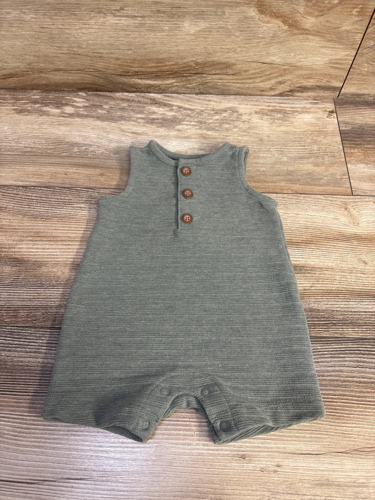 Front view of a green Just One You Henley tank romper showcasing a three-button placket, ribbed texture, and snap closures on wood grain