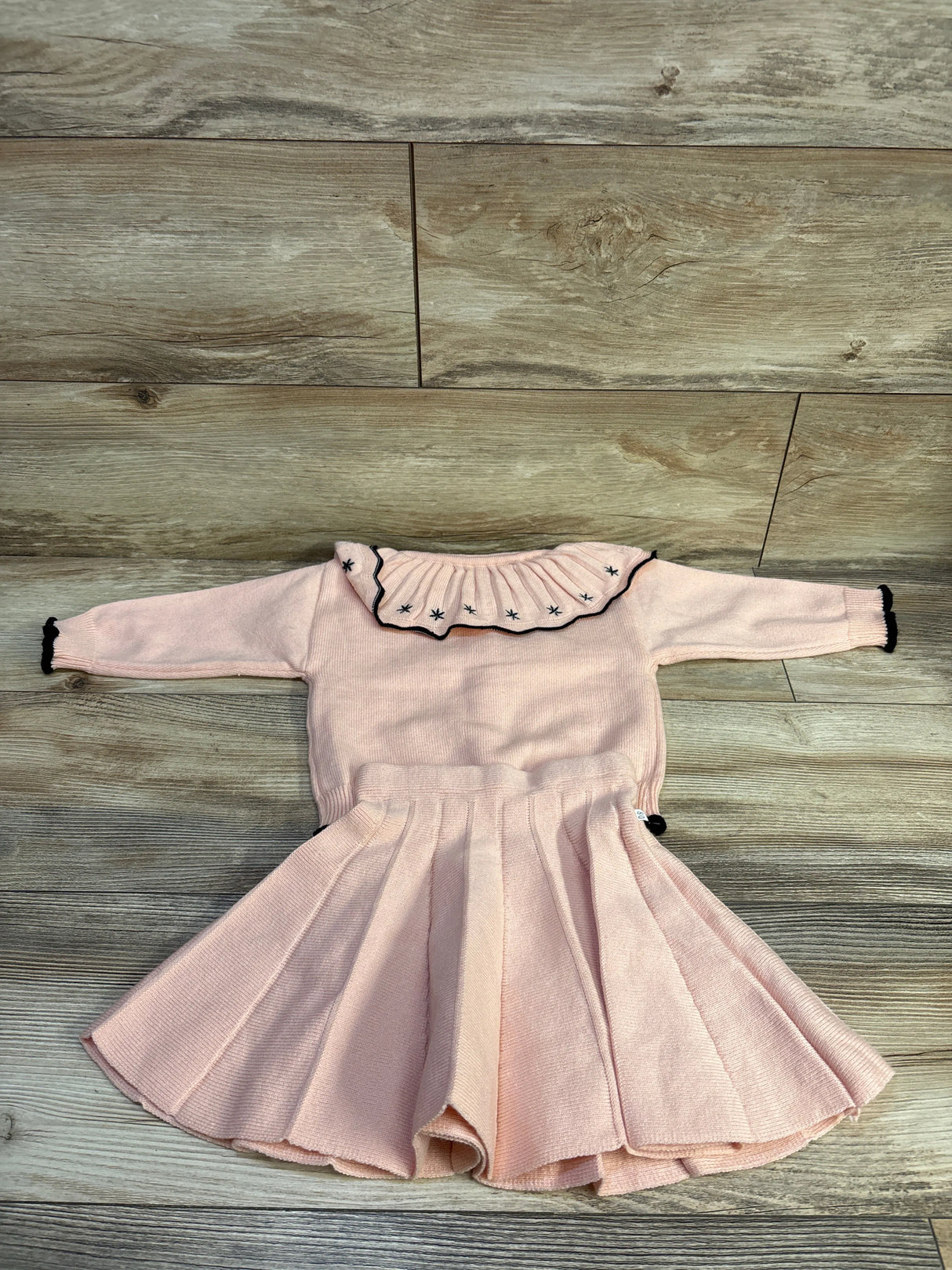 Flat lay of pink 2pc knit cardigan and pleated skirt set with ruffled collar, black trim, star accents on wood floor