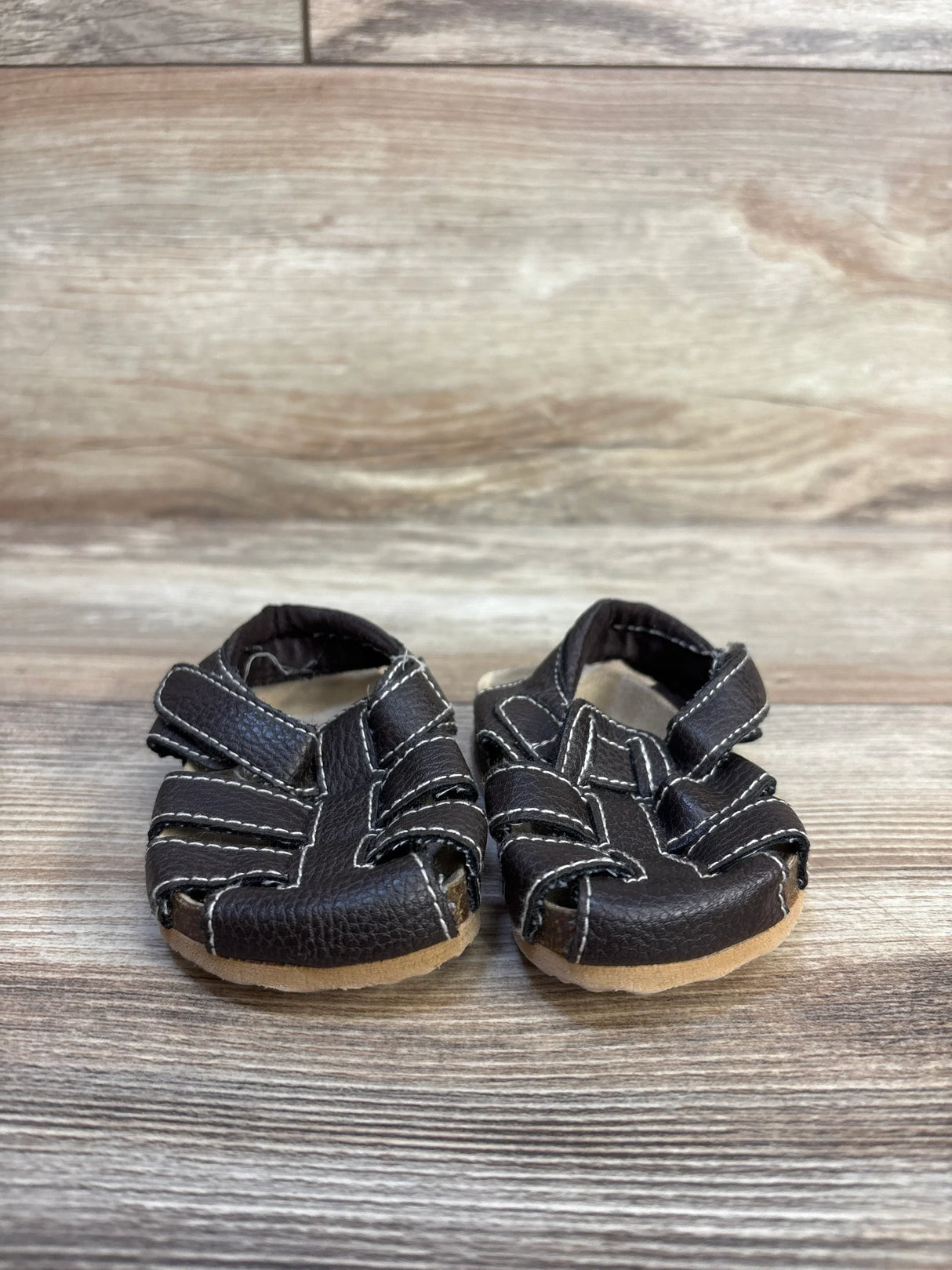Front view of Childrens Place leather fisherman sandals in dark brown with white contrast stitching on wood plank floor