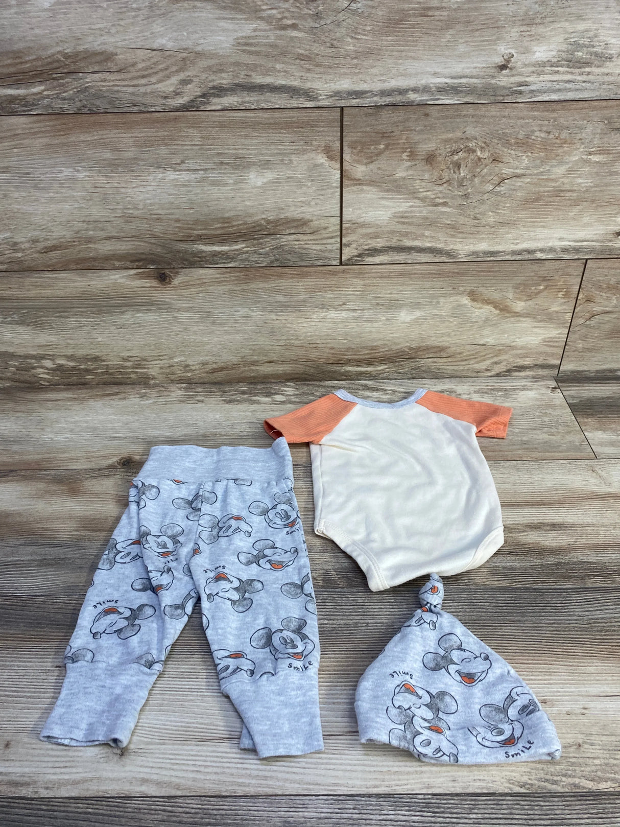 Flat lay of newborn Disney 3pc Mickey set: cream bodysuit with orange sleeves, gray Mickey pants and knotted hat on wood