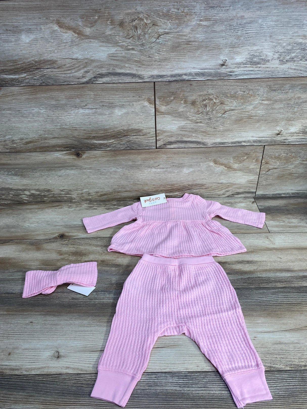 Flat lay of NWT Cat and Jack 3pc waffle knit set in pink showcasing peplum top ribbed pants and headband on wood plank floor