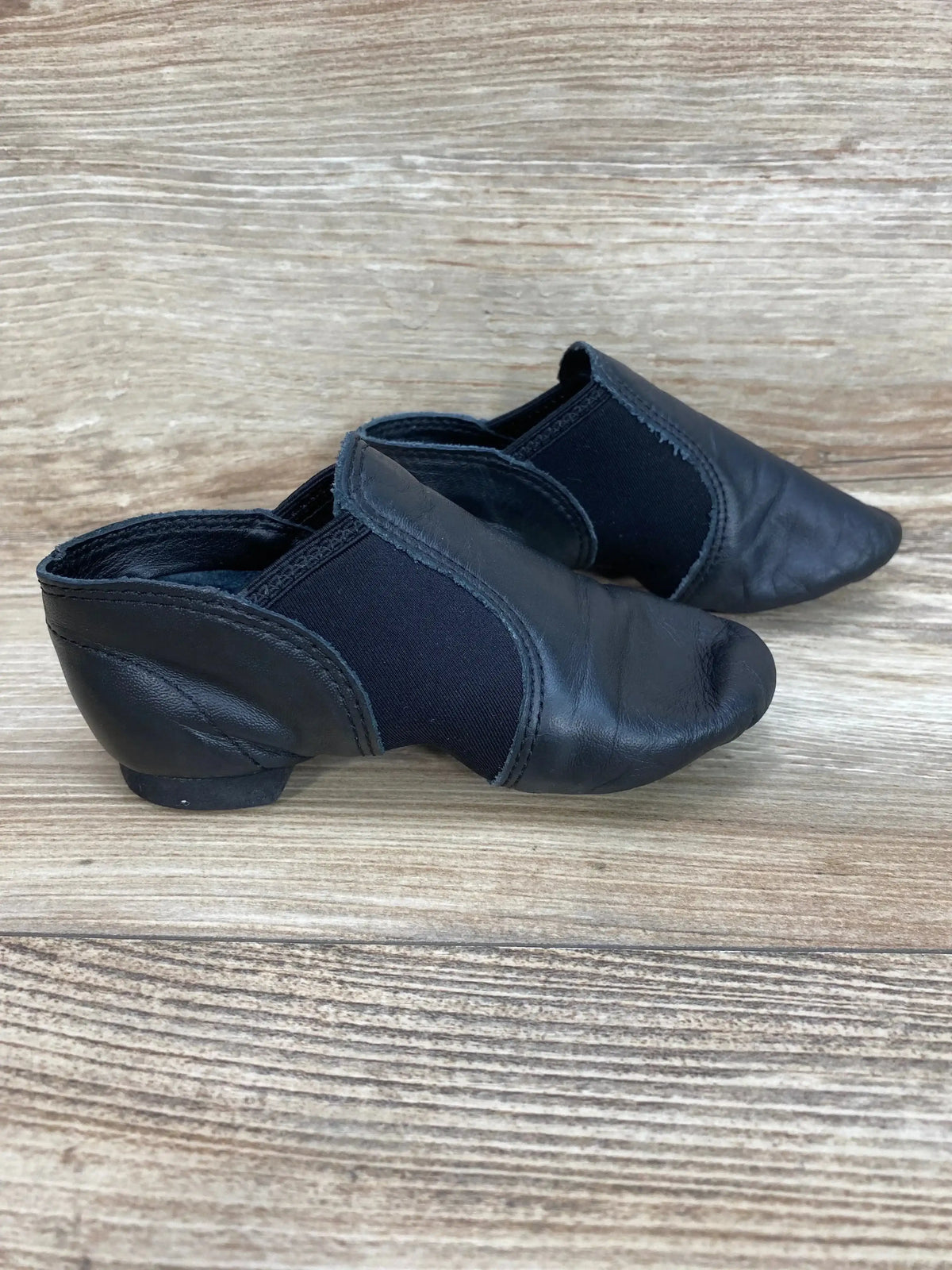 Side view of Capezio black jazz slip-on shoes showing leather upper, elastic side panels and low heel on wood background