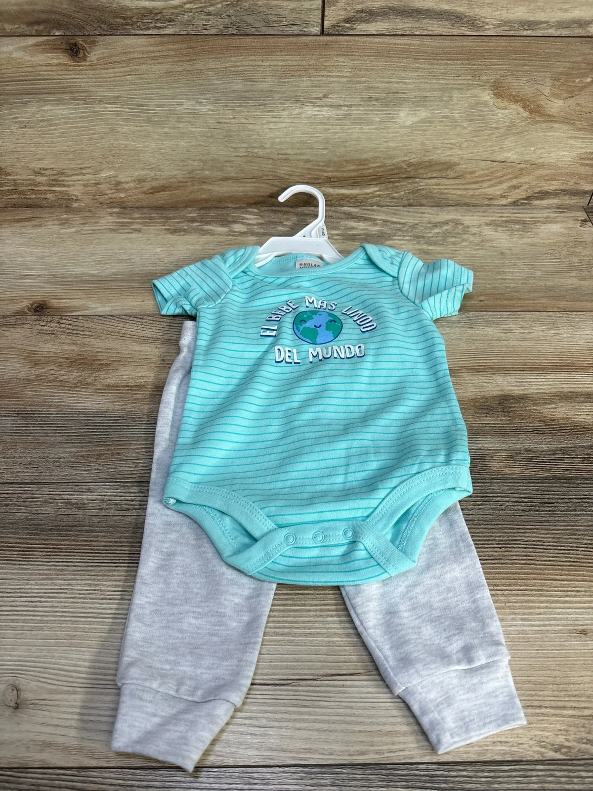 Top-down view of teal striped baby bodysuit with ‘El bebe mas lindo del mundo’ globe graphic and gray pants on wood