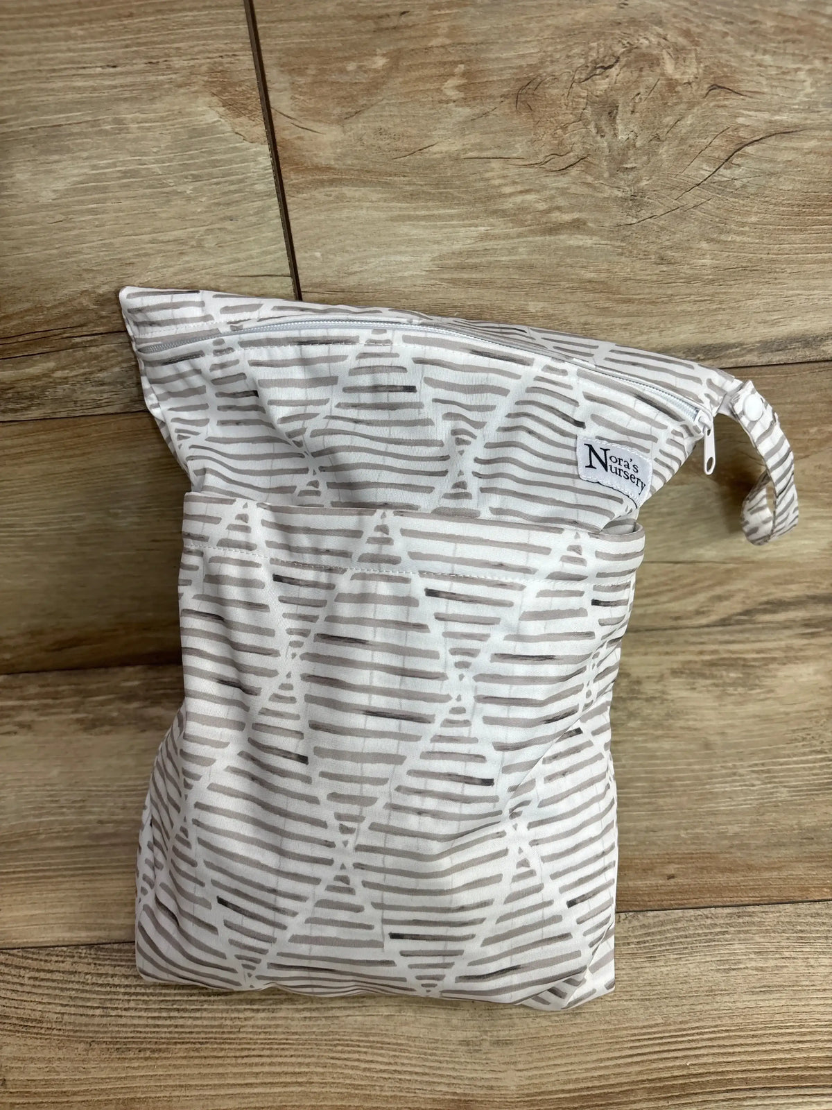 Front view of Nora’s Nursery wet bag showcasing gray and white stripes, zipper closure, and hanging loop on wood surface