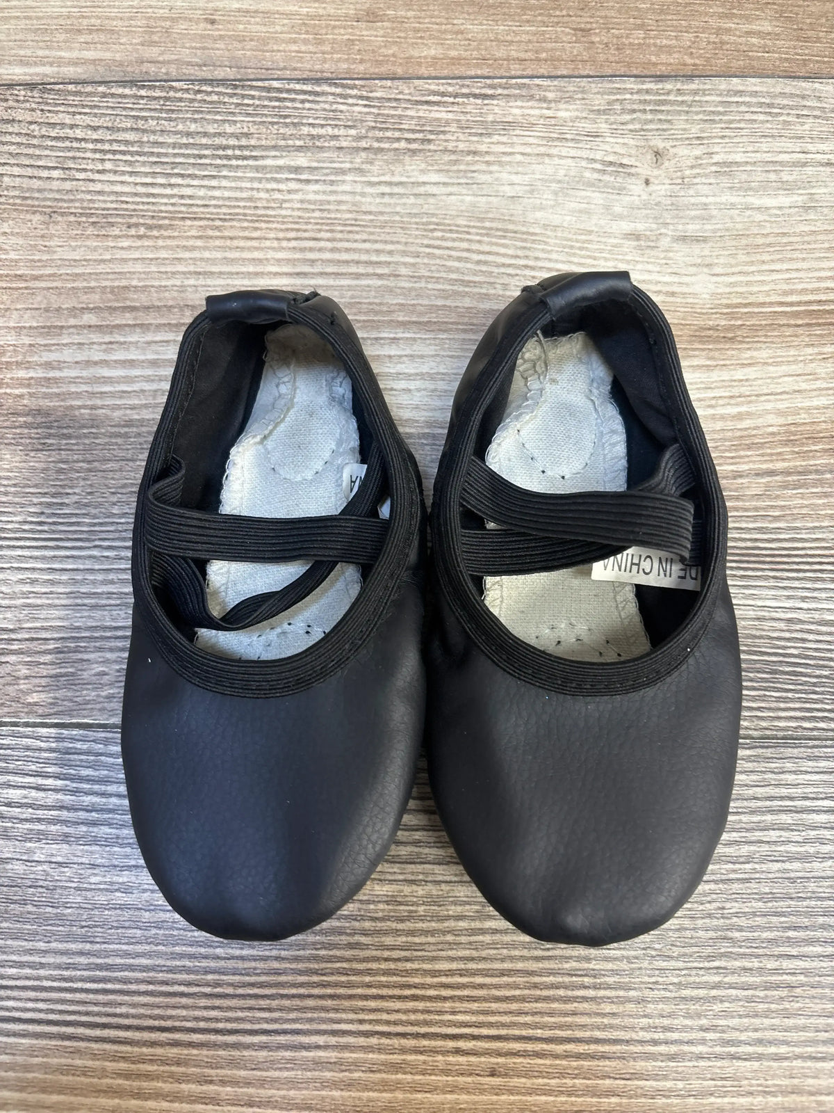 Black toddler ballet slippers with elastic straps on wood floor, gently used kids shoes