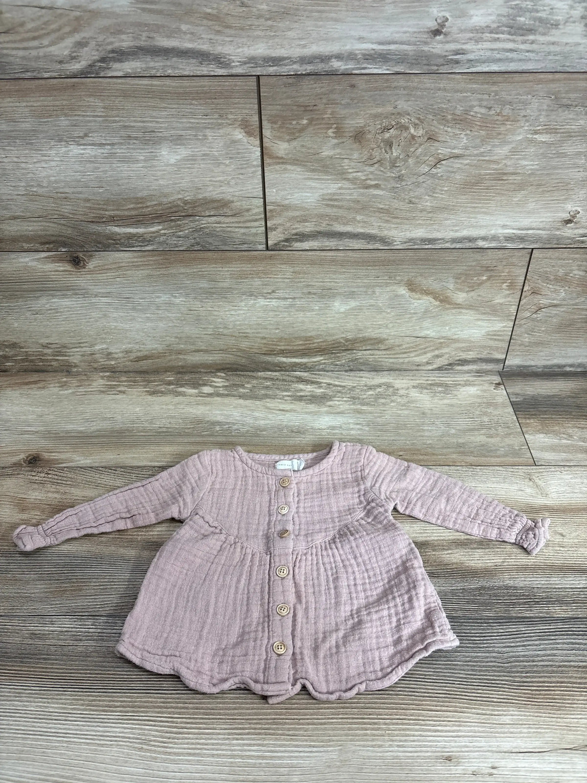 Flat lay of Jamie Kay Lila muslin blouse in rose smoke showing front wooden buttons, long sleeves, gathered waist and scalloped hem on wood floor