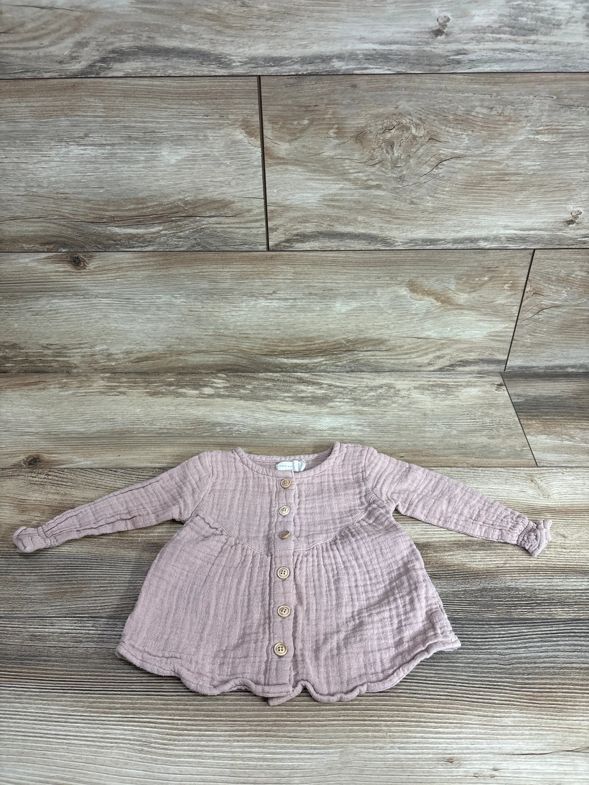 Flat lay of Jamie Kay Lila muslin blouse in rose smoke showing front wooden buttons, long sleeves, gathered waist and scalloped hem on wood floor