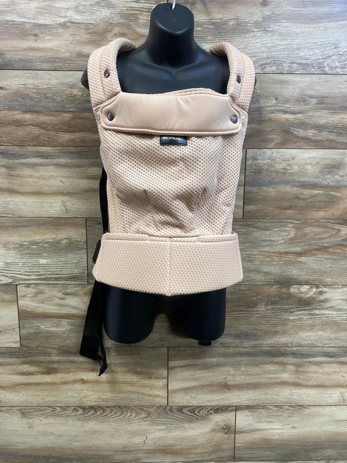 Front of Momcozy PureHug baby carrier in beige mesh with padded straps and wide waist belt on mannequin, wood wall background