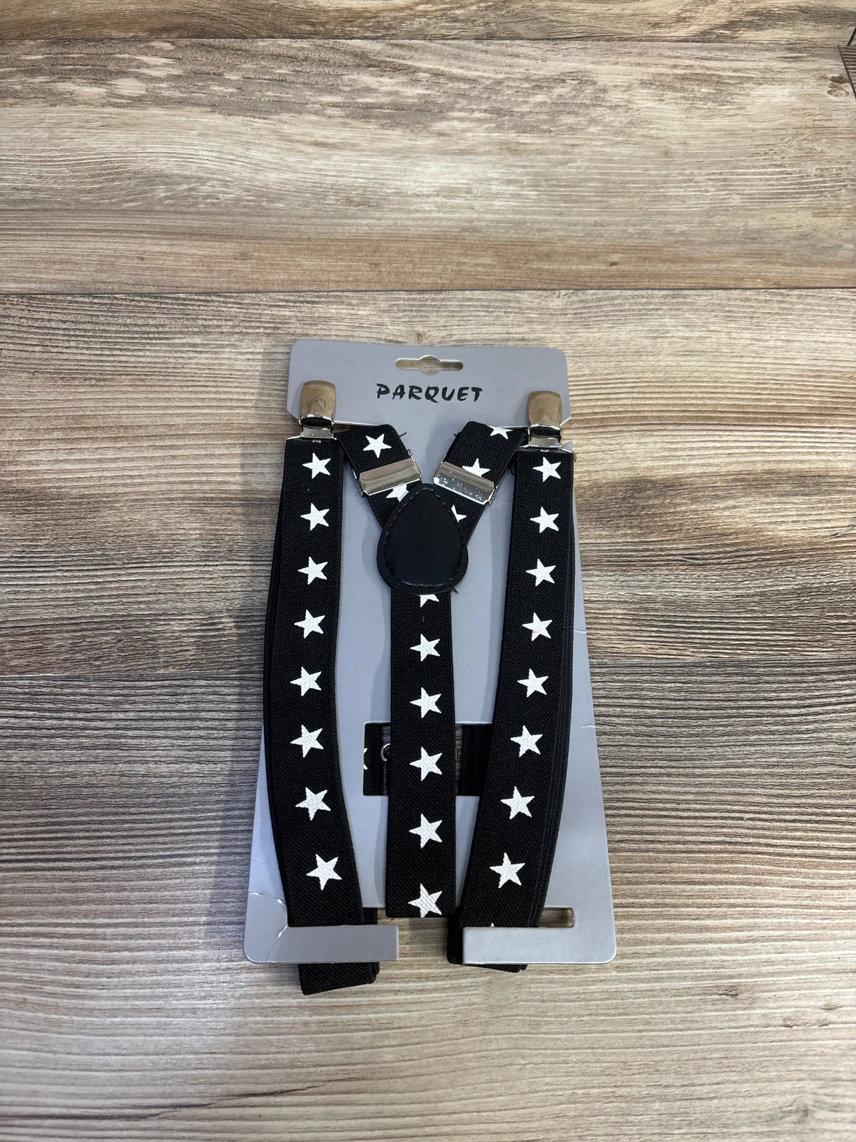 Front view of Parquet star print suspenders showcasing black straps with white stars, metal clips, and a wood-grain background